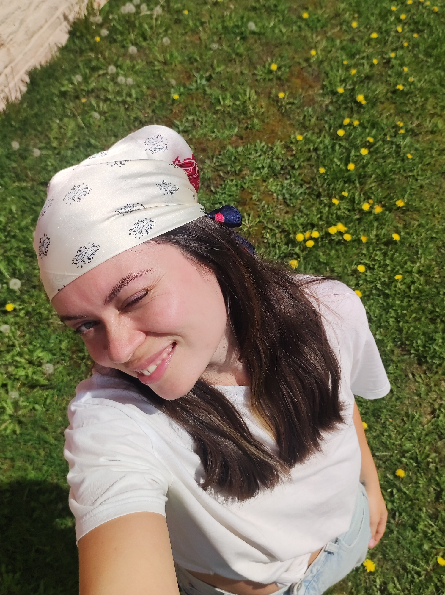 A young woman is seen in an overhead, candid shot taken in daylight outdoors. She has fair skin, dark hair, and wears a white t-shirt, light blue denim shorts, and a patterned headscarf. She is winking at the camera and smiling. The background consists of green grass dotted with yellow dandelions. The location is likely not Фоки, Russia, but a similar sunny, grassy area. There are no discernible city-specific details present in the image.