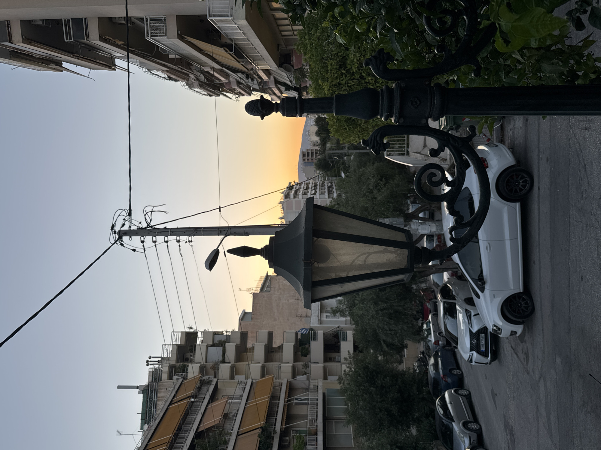 The image captures an urban street scene in Athens, Greece, during twilight. The foreground is dominated by a decorative black lamppost with ornate scrollwork, partially obscuring a white Volkswagen Polo. In the midground, several cars are parked along the street, including a silver hatchback, a dark blue sedan, and a white Lexus. The street curves gently uphill, leading the eye towards the background where apartment buildings line both sides. These buildings feature balconies with awnings and are interspersed with trees. Power lines crisscross the sky, and a utility pole is visible. The sky displays a gradient of pale blue to soft orange, indicating the setting sun, with a faint silhouette of hills on the horizon. The overall impression is a residential street in Athens as daylight fades.