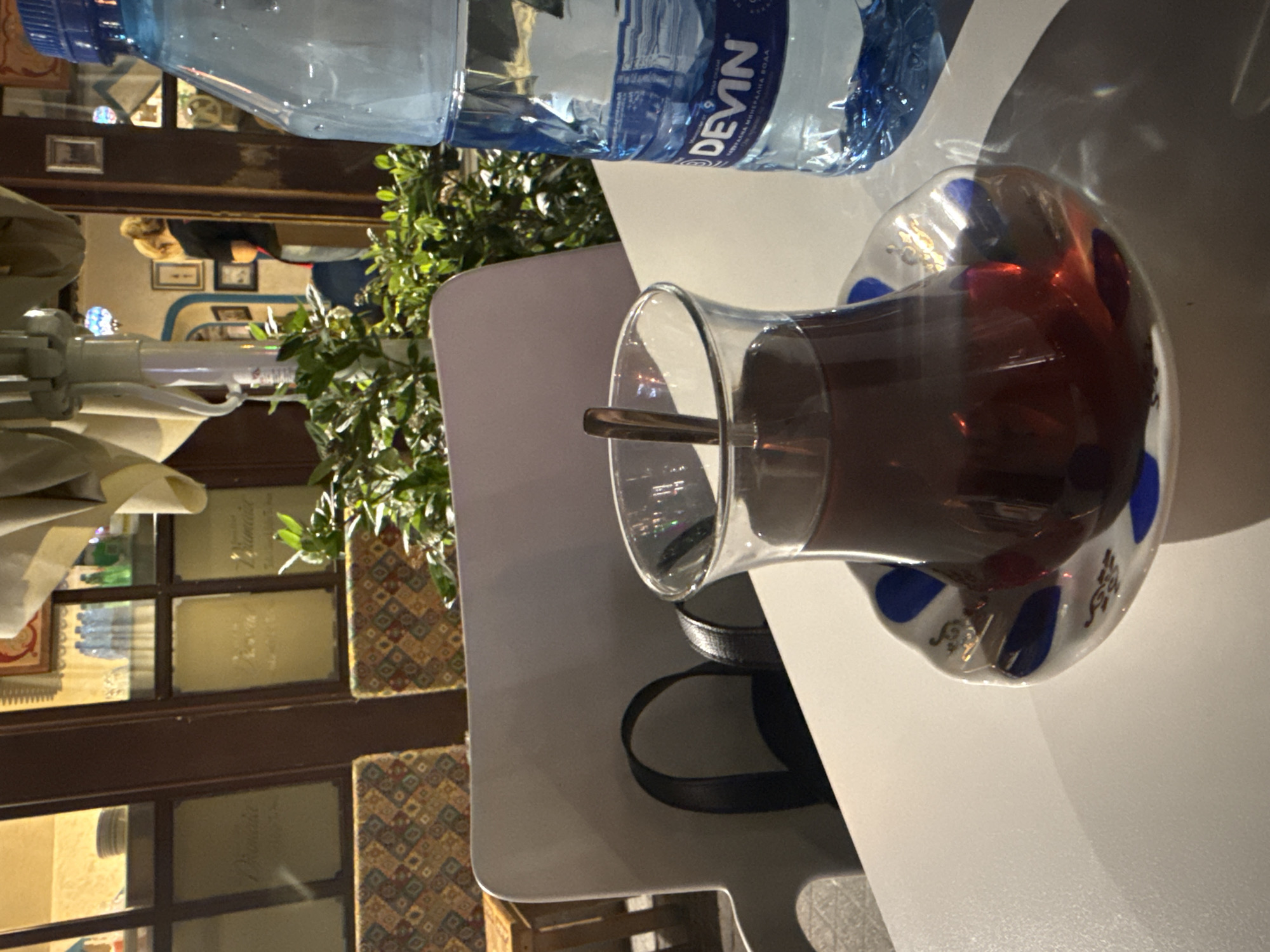 The image depicts a glass of tea on a white table, with a bottle of "Devin" water nearby, likely at a restaurant in Plovdiv, Bulgaria. Through the window, a person with blond hair can be seen, as well as details of the building interior. A decorative plant sits adjacent to the tea. A chair with patterned fabric is also visible. A spoon rests inside the glass of tea.