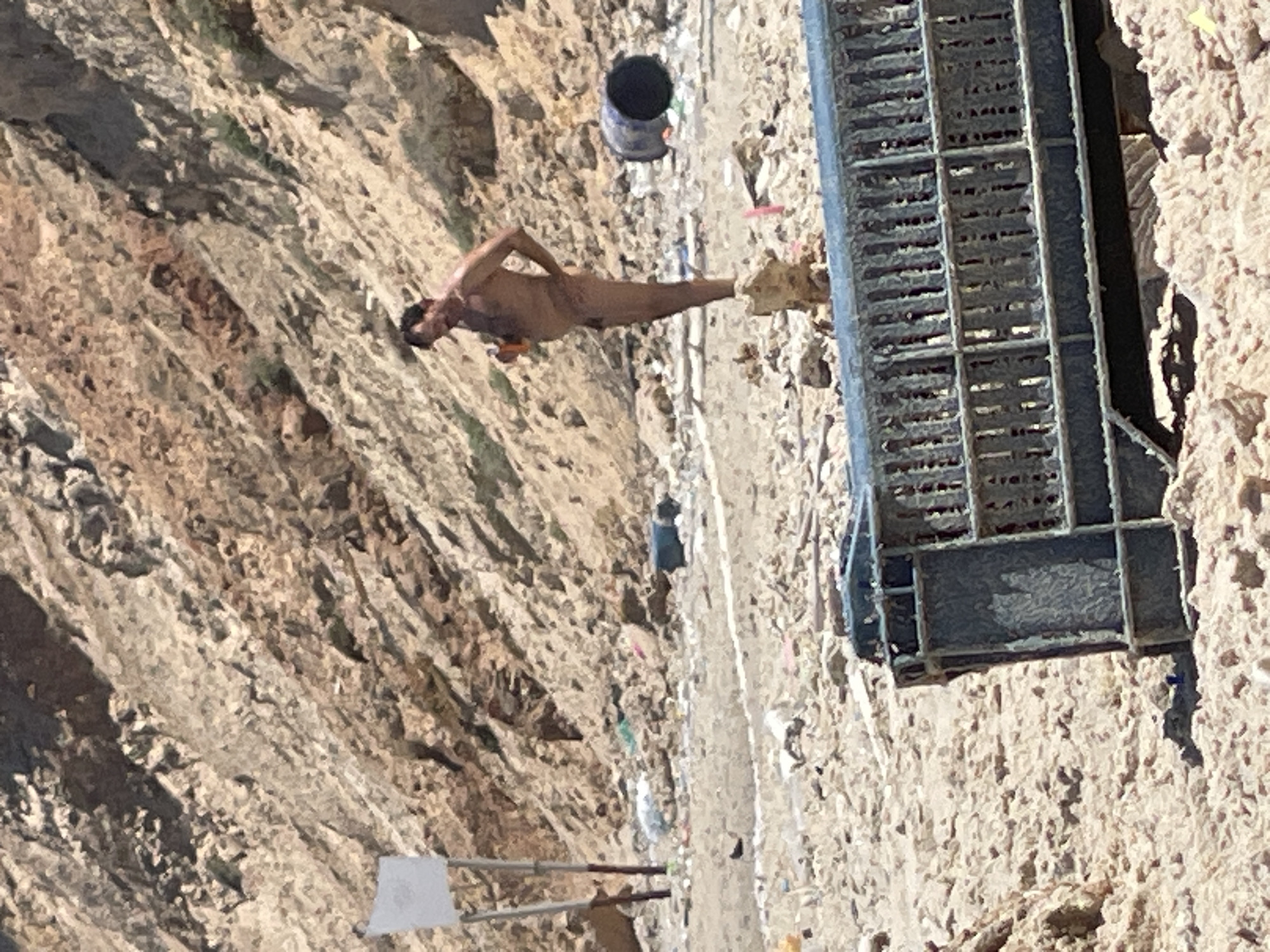 The image depicts a man standing nude on a beach, holding an orange object in his right hand. He is facing to the left of the frame and his left hand is on his hip. The background consists of a rocky hillside and a beach littered with various debris, including a large black barrel and what appears to be a discarded metal cage in the foreground. A white sign on a pole is visible to the left of the man. The location is the coast of Ga'ash, Israel.