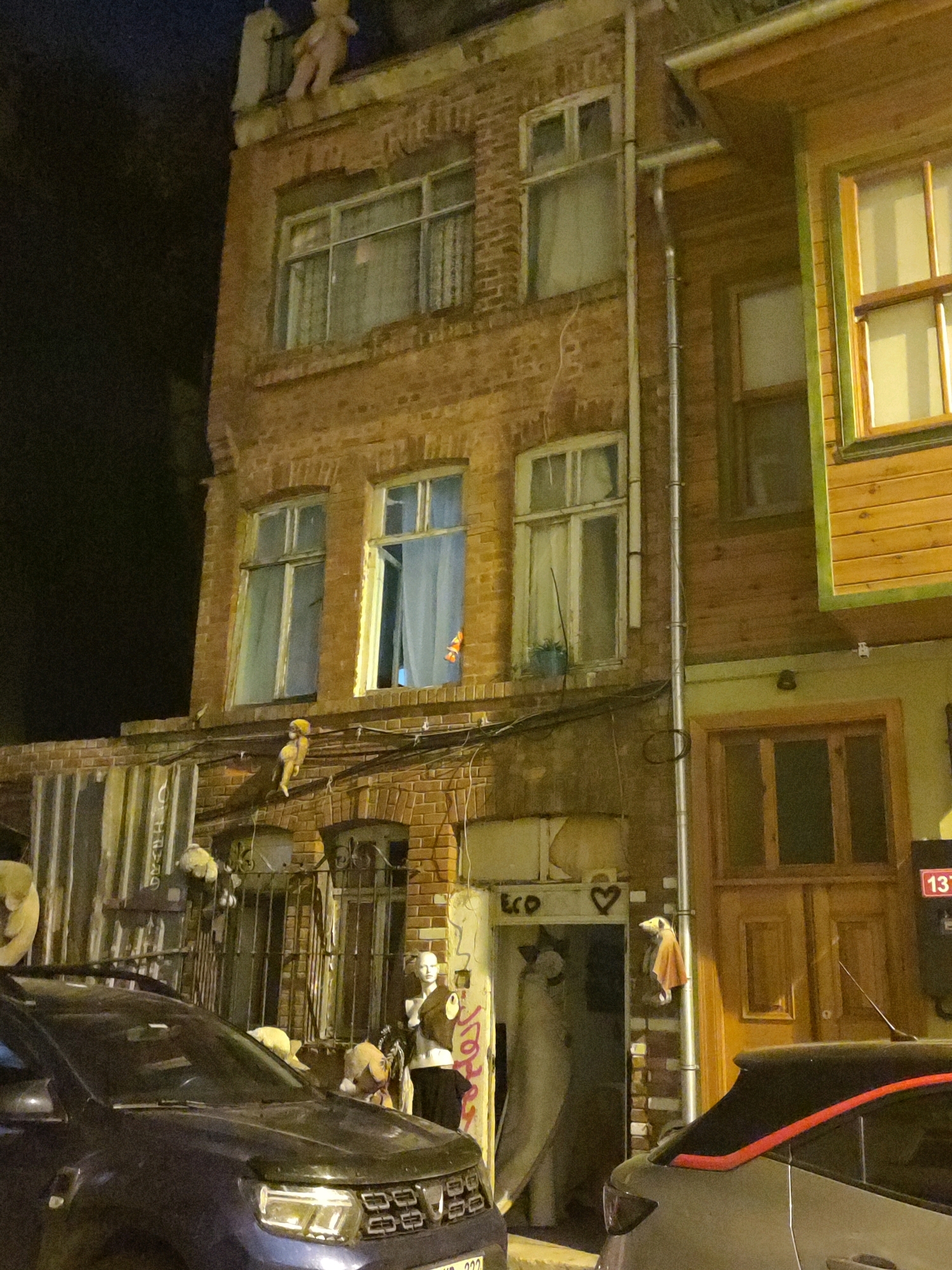 The image depicts a brick building in Kadıköy, Turkey, at night. The building has multiple windows, some with curtains visible. Teddy bears and other stuffed animals are placed on the roof and balconies of the building. A dark-colored SUV is parked in front of the building. To the right of the brick building is a wooden-clad structure with windows. The scene appears to be a street with some artistic or decorative elements.