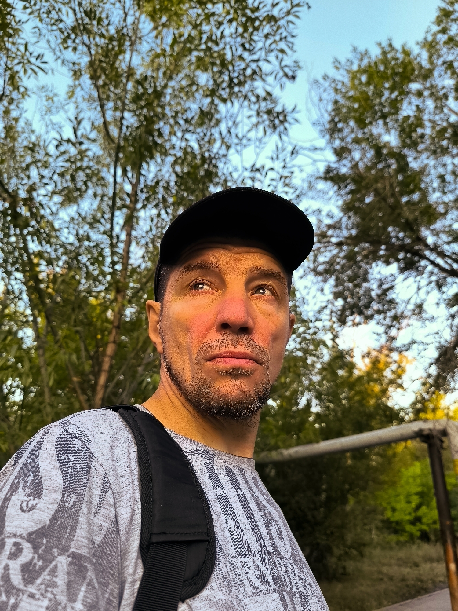 A medium shot shows a man from the chest up, looking up and to the right. He is wearing a grey t-shirt with a graphic pattern and a black baseball cap. A black strap from a backpack is visible across his shoulder and chest. The background consists of trees with green and yellow foliage and a blue sky, suggesting an outdoor setting. The lighting indicates it is likely late afternoon or early morning, with warm light illuminating the man's face. The presence of a metal frame structure in the background, partially visible, suggests a recreational area. The setting is not specifically identifiable as being in Qarağandı city administration, Kazakhstan.