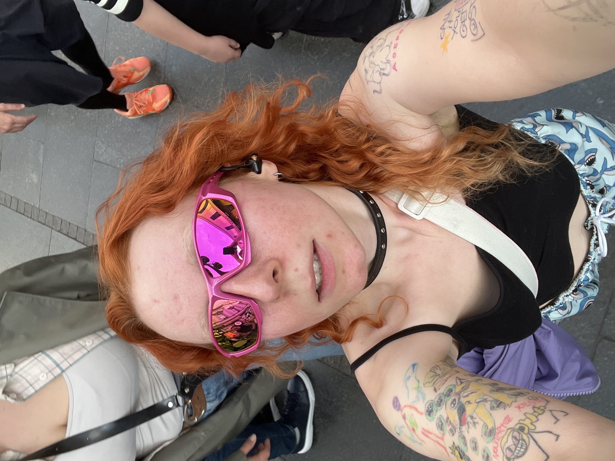 A selfie taken at ground level shows a young person with red, curly hair and pink sunglasses, a black studded collar, and several arm tattoos. The subject is wearing a black tank top with white straps and a drawstring bag with a shark pattern. In the background, parts of other people are visible. One person is wearing orange sneakers. The location is Belgrade, Serbia, as evidenced by the reflections in the sunglasses.