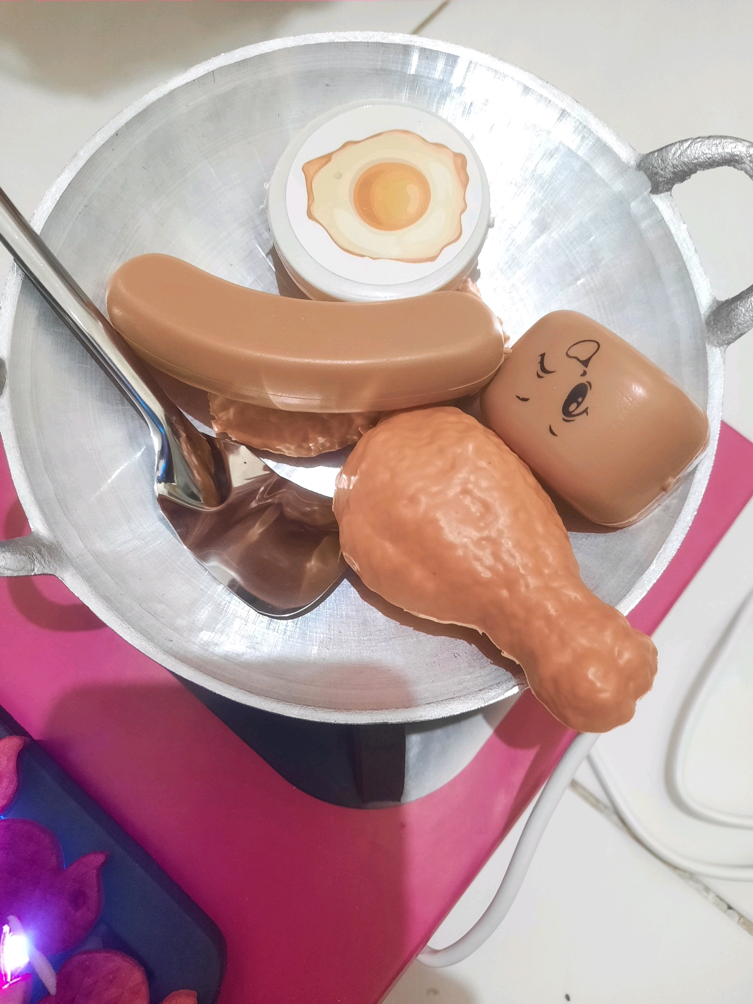 The image shows a toy frying pan containing toy food items: a fried egg, a sausage, a chicken drumstick, and a smiling cup with a face. A metal spoon is also inside the pan. The frying pan is placed on a bright pink surface, which appears to be a mat or a table runner. A white cable runs along the white surface next to the pink mat. The background is a white tiled surface and a wall. The scene suggests a playful or educational setting, possibly a child's play area. The image does not provide any specific information that can be used to determine its location in North Jakarta, Indonesia.