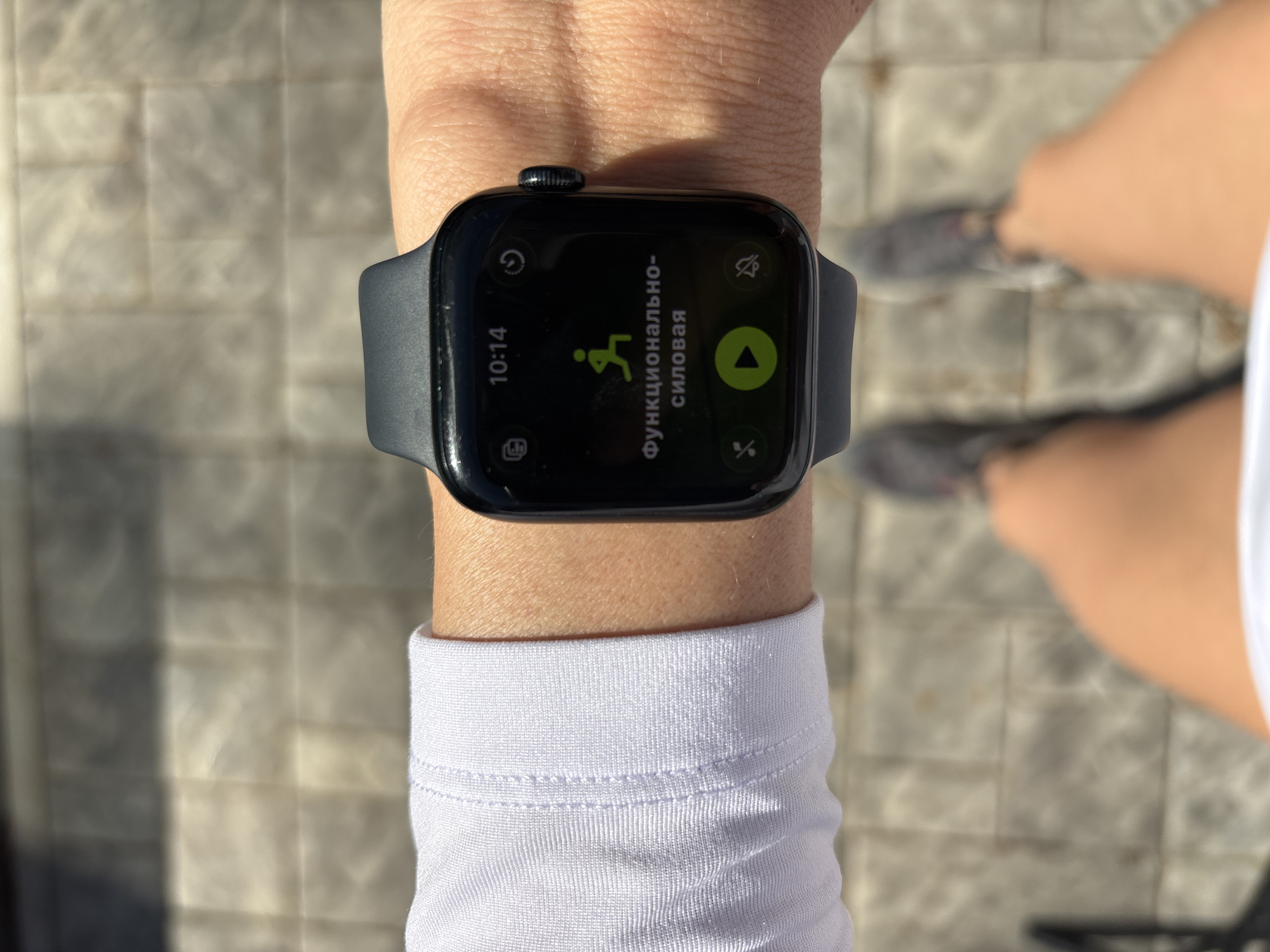 A person is wearing a black Apple Watch on their left wrist. The watch face displays "10:14" and Cyrillic text that translates to "Functional Power" along with a stylized icon of a person running. The person is wearing a white long-sleeved shirt. The background shows paving stones and a blurred view of legs and feet wearing gray shoes. It's impossible to verify the location without additional information.
