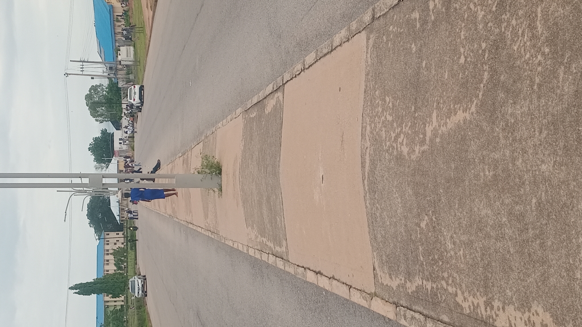 The image shows a street scene in Jalingo, Nigeria, with a person in a blue dress standing on the sidewalk. The street is paved and bordered by a sidewalk with a textured surface. In the background, there are buildings, trees, and a cloudy sky. Several people are visible along the street, some sitting and others walking. A white vehicle is parked on the side of the road. Streetlights are present on the sidewalk. The image appears to have been taken during the daytime. The overall scene depicts a public area with everyday activities.