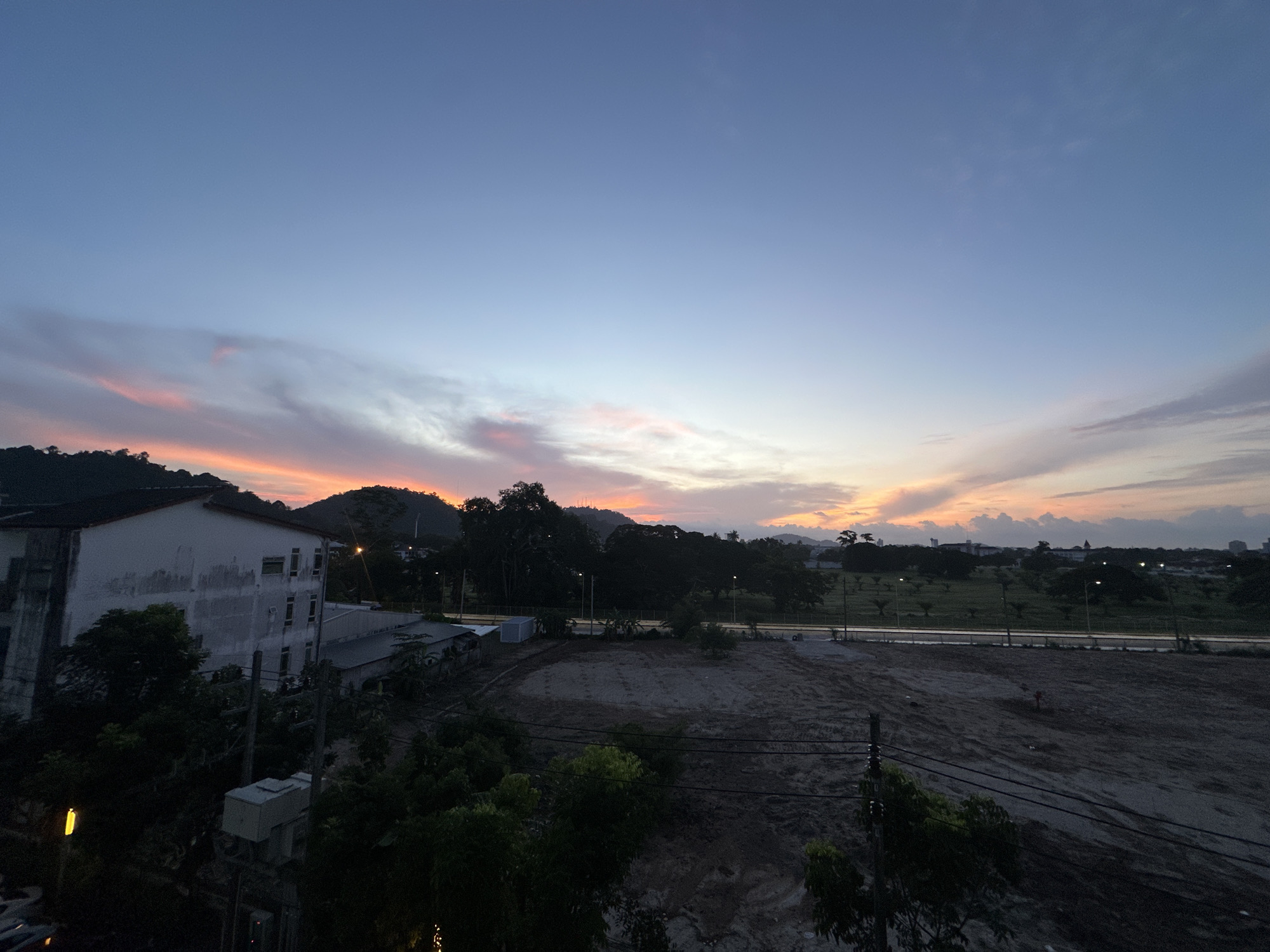 A wide-angle shot captures a twilight sky over a semi-urban landscape. The sky transitions from a pale blue at the top to hues of pink, orange, and yellow closer to the horizon, with wispy clouds streaked across. In the foreground, a dark, mostly cleared plot of land is visible, with scattered debris and tire tracks suggesting recent activity. To the left, a weathered, multi-story building with a white facade and dark roof stands partially in shadow. Behind the building and plot of land, a line of dark trees forms a backdrop, beyond which more distant buildings and a treeline can be seen against the fading light. Power lines and utility poles are present in the mid-ground and foreground, with a single yellow light fixture visible on a pole in the lower left. The overall impression is a developing or transitional area within a city, as day turns to night.