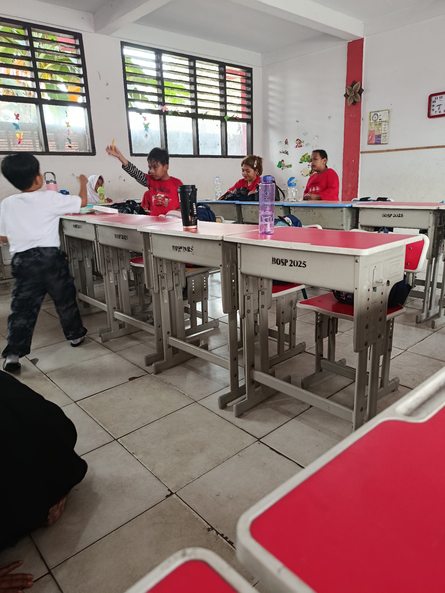 In a classroom in Balaraja, Indonesia, a group of children are seated at desks, engaged in what appears to be a lesson. One boy in a red shirt, seated in the foreground, is actively participating, holding a yellow pencil aloft. To his left, another child wearing a white hijab is also looking towards the front of the classroom. In the background, two other children in red shirts are seated at their desks, with one appearing to look out of frame. A boy in a white shirt and camouflage pants is standing and facing away from the camera, also looking towards the front. The classroom is equipped with rows of desks with red tabletops and grey metal frames. The walls are painted white, with colorful decorations and a clock visible. Sunlight streams in through windows with horizontal blinds, illuminating the scene.