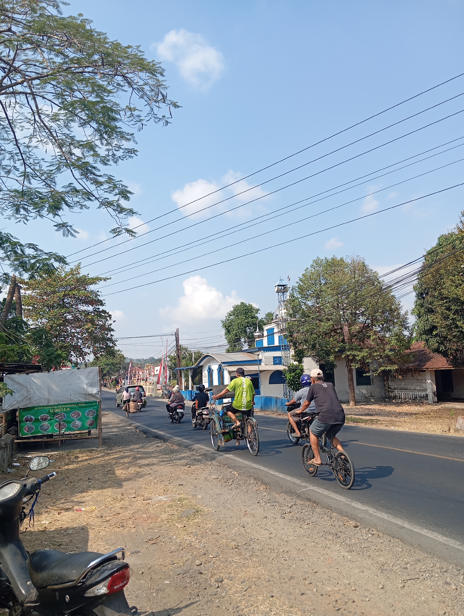 The image depicts a street scene in Sokaraja, Indonesia, on a bright, sunny day. A paved road with a white line marking runs through the center of the frame, with a gravelly shoulder on the left. Several people are engaged in various modes of transportation. In the foreground on the left, a black scooter is parked. On the road, a person is riding a small bicycle. Further down the road, a motorcycle carries two passengers, and another motorcycle carries a single rider. Two other individuals are riding bicycles, one of which appears to be a tricycle. On the left side of the road, a stall with a green tarp displays food advertisements. Buildings line the right side of the road, including a white structure with blue accents and a mosque with a tall minaret. Lush green trees and power lines are visible throughout the scene, with a clear blue sky dotted with white clouds above.