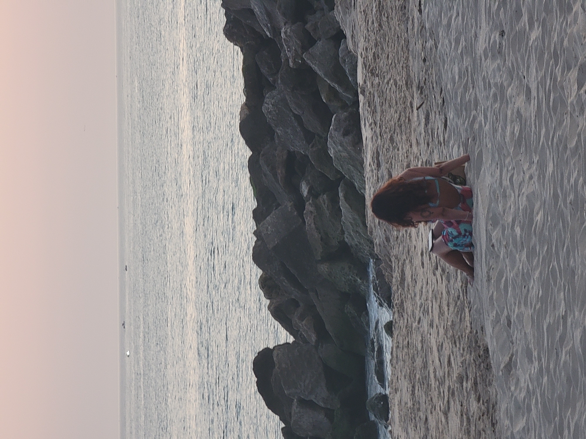 Two individuals are seated on a sandy beach, with a rocky breakwater in the background. The ocean stretches out to the horizon, where several small boats are visible. The sky is a soft pinkish hue, suggesting either sunrise or sunset. The individual in the foreground, closer to the viewer, is partially visible from the back, with dark, long hair. They are wearing patterned shorts and a bikini top. Their legs are bent, and they appear to be sitting upright, possibly looking at something on their lap. A second individual is partially visible behind and slightly below the first, with their legs extended. They are wearing a patterned garment that covers their lower body and appears to be leaning against a bag. The beach is composed of fine, light-colored sand, with ripples and shadows indicating the texture. The rocks of the breakwater are dark and irregular in shape, with some appearing to be covered in moss or seaweed. The water is calm, with gentle waves lapping at the shore. The scene is peaceful and serene, conveying a sense of relaxation and quiet enjoyment of the coastal environment. The location is the beach in Quarteira, Portugal, known for its extensive sandy shores and coastal fortifications.