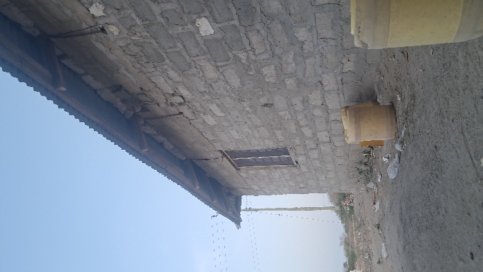 The scene depicts the exterior of a building located in Walu, Kenya. The structure is constructed from unpainted grey concrete blocks with visible mortar lines and some surface irregularities. A corrugated metal roof with a visible wooden overhang extends from the building. Electrical wiring is affixed to the upper portion of the exterior wall, below the roofline. A single, dark-framed window with vertical security bars is present on the side of the building.
The ground in the foreground and adjacent to the building is unpaved dirt, appearing dry and uneven, with scattered white plastic debris and other litter. Two large, light yellow cylindrical containers are visible; one is partially in the foreground on the right, and the other stands upright against the building's wall.
To the left of the building, a utility pole with several overhead wires extends into the distance. The background consists of sparse vegetation under a hazy, light blue sky. No individuals are present in the image.