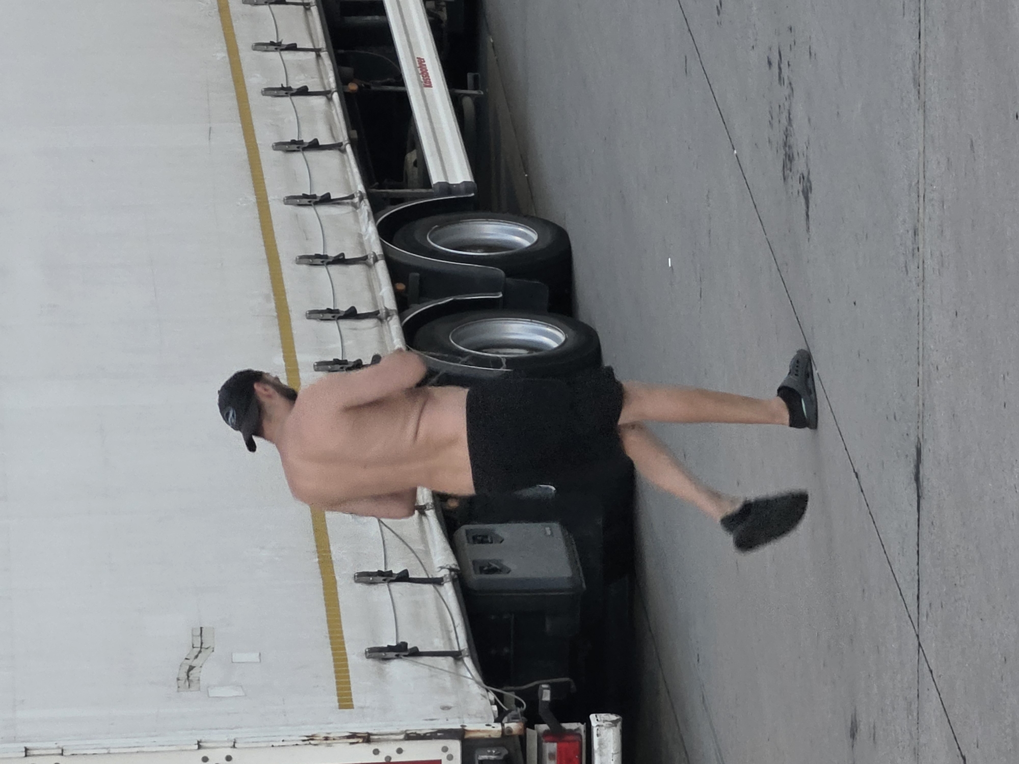 A shirtless person wearing a black baseball cap and black shorts is walking away from the camera in front of a white semi-trailer truck. The truck has a yellow stripe running horizontally across its side. The person is on a gray, paved surface that appears to be an industrial area or parking lot. The scene is outdoors. There is no indication of this image being located in Frankfurt, Germany.