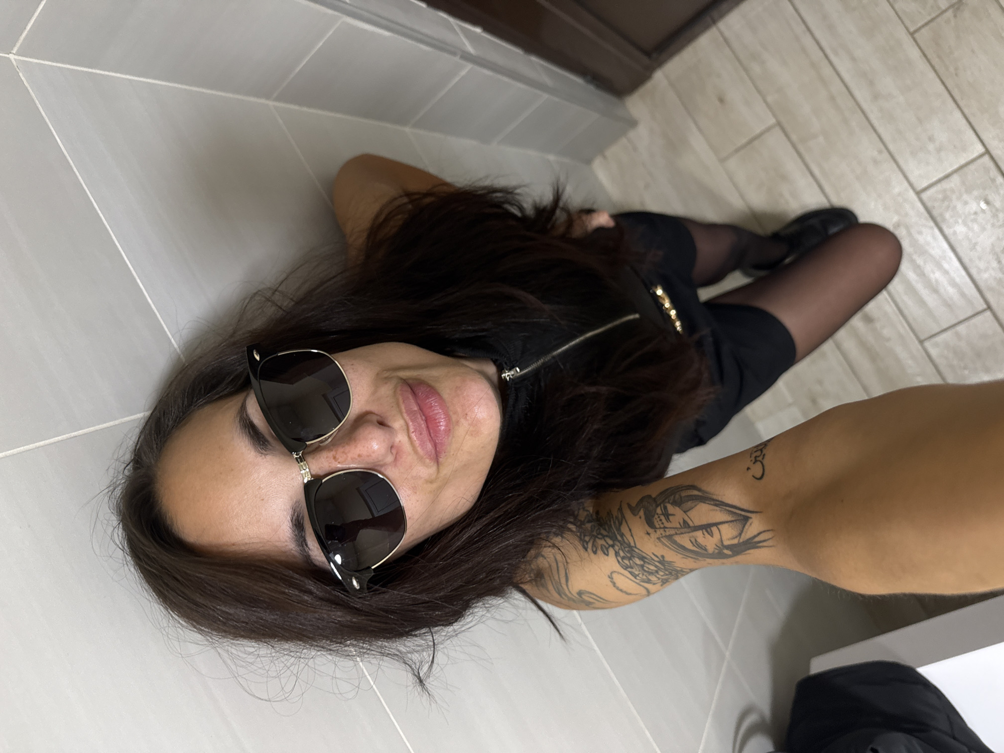 A woman is lying on a tiled floor. She is wearing sunglasses, a black dress, and black tights with shoes. She has long brown hair and tattoos on her left arm. The location appears to be indoors, possibly a bathroom, given the tile walls and floor. There is no information to determine if the image was taken in Byala Slatina, Bulgaria.