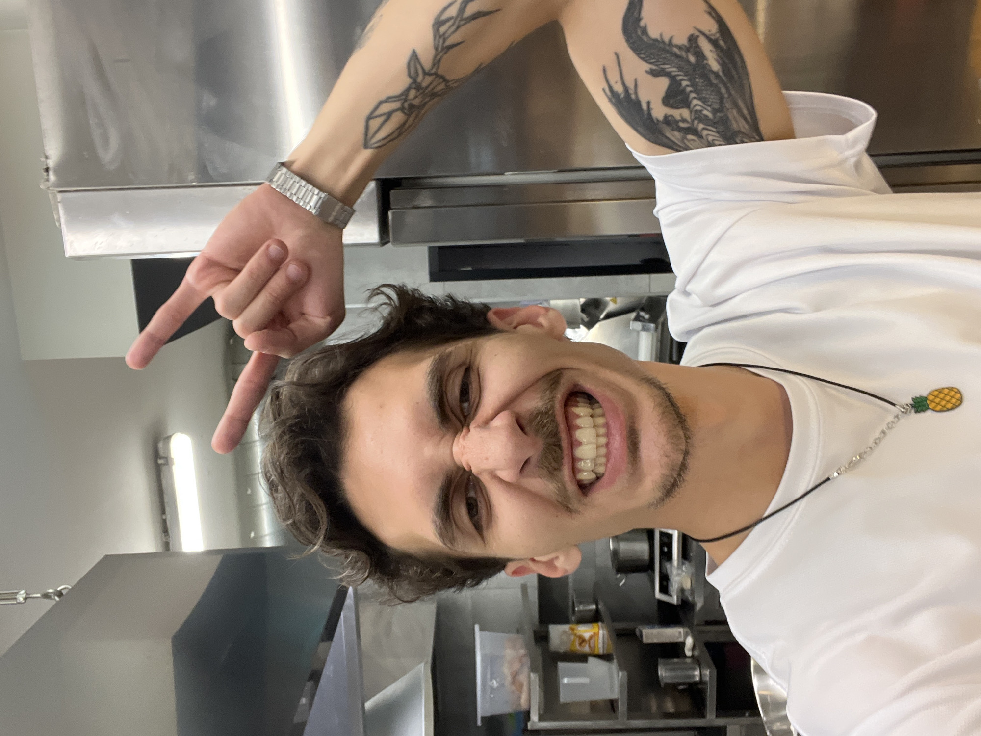 A single male individual is depicted in a selfie photograph taken within a commercial kitchen environment, located in Budva, Montenegro. The subject, a young adult with dark, curly hair, a mustache, and light stubble, is shown from the chest up, facing the camera directly. His facial expression is exaggerated, featuring a scrunched nose and a wide, open-mouthed grin revealing his teeth. His left hand is raised, forming a "peace" sign with the index and middle fingers, positioned near the top of his head. He wears a white short-sleeved t-shirt, a black cord necklace with a silver chain segment and a yellow-green pineapple pendant, and a silver-colored metal link wristwatch on his left wrist. Visible tattoos on his left arm include an abstract or geometric design on the forearm and a dark, winged creature or stylized design on the bicep/upper arm.
The background consists of a functional kitchen space, characterized by stainless steel surfaces. A stainless steel ventilation hood is visible to the upper left. To the right, a large stainless steel refrigeration unit or oven is present. In the mid-ground left, shelving holds various kitchen items, including clear plastic containers with contents, smaller cups, bottles, and silver pots. The ceiling is white, with a visible fluorescent light fixture providing illumination.