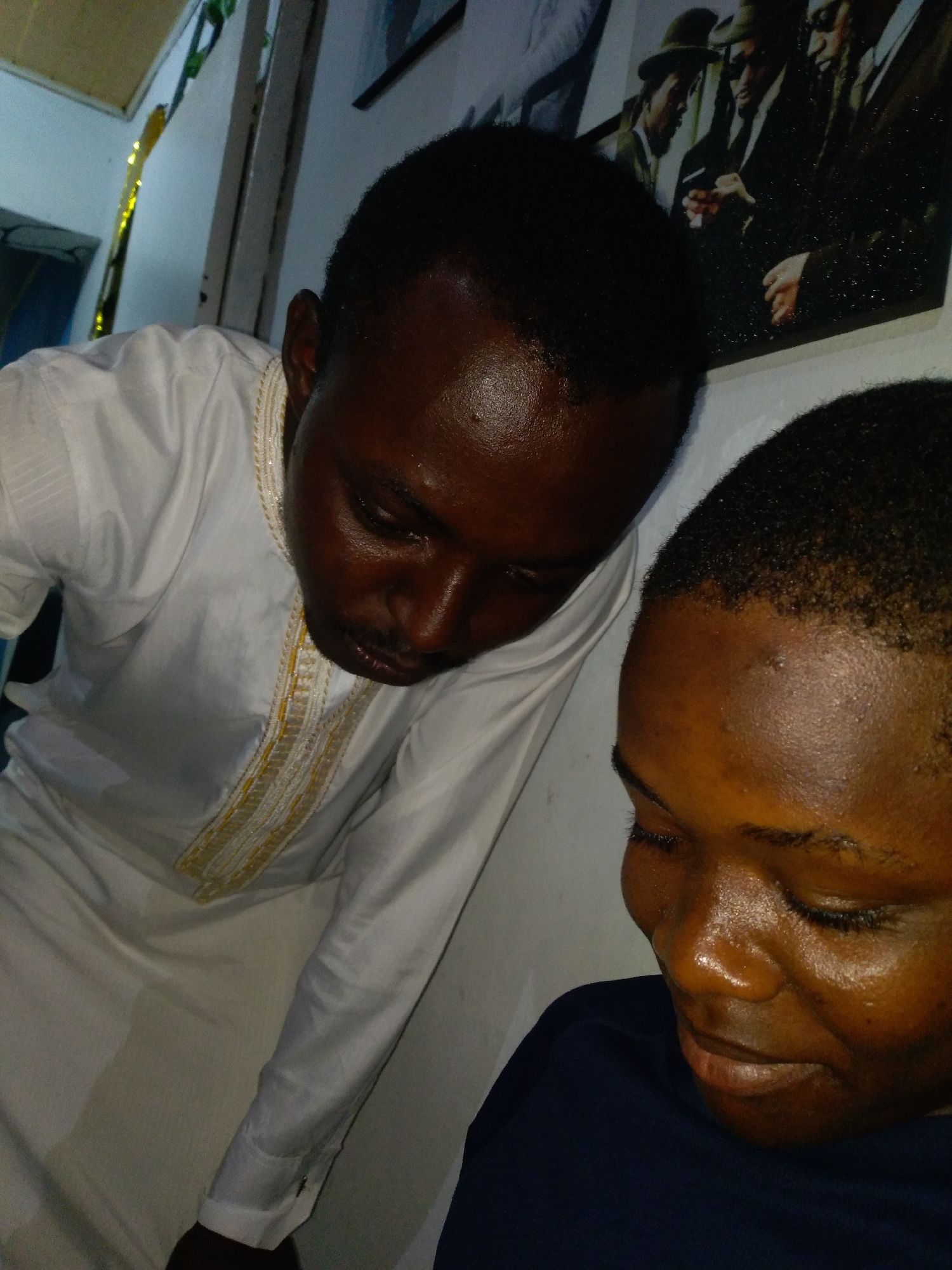 Two individuals with dark skin tones are depicted indoors in Unknown, Unknown. The person on the left, an adult male, is positioned vertically, wearing a white long-sleeved garment with prominent gold and white embroidery detailing the placket. He is looking downwards and to the right, with a visible mustache and slight stubble. His skin appears reflective, possibly due to oil or sweat. The person on the right, also an adult male, is partially visible from the head and right shoulder, wearing a dark blue top. His head is tilted downwards and slightly to the left, with eyes either closed or cast very low, and his facial expression appears serene or focused. His skin also exhibits a reflective quality. The two individuals are in close proximity, suggesting an interaction or shared focus. In the background, a framed picture is mounted on a light-colored wall, depicting several men in vintage attire, including fedoras and suits, appearing to interact. To the far left, a strip of shimmering gold decoration is visible, alongside architectural details of a ceiling and wall. The lighting in the scene is subdued.