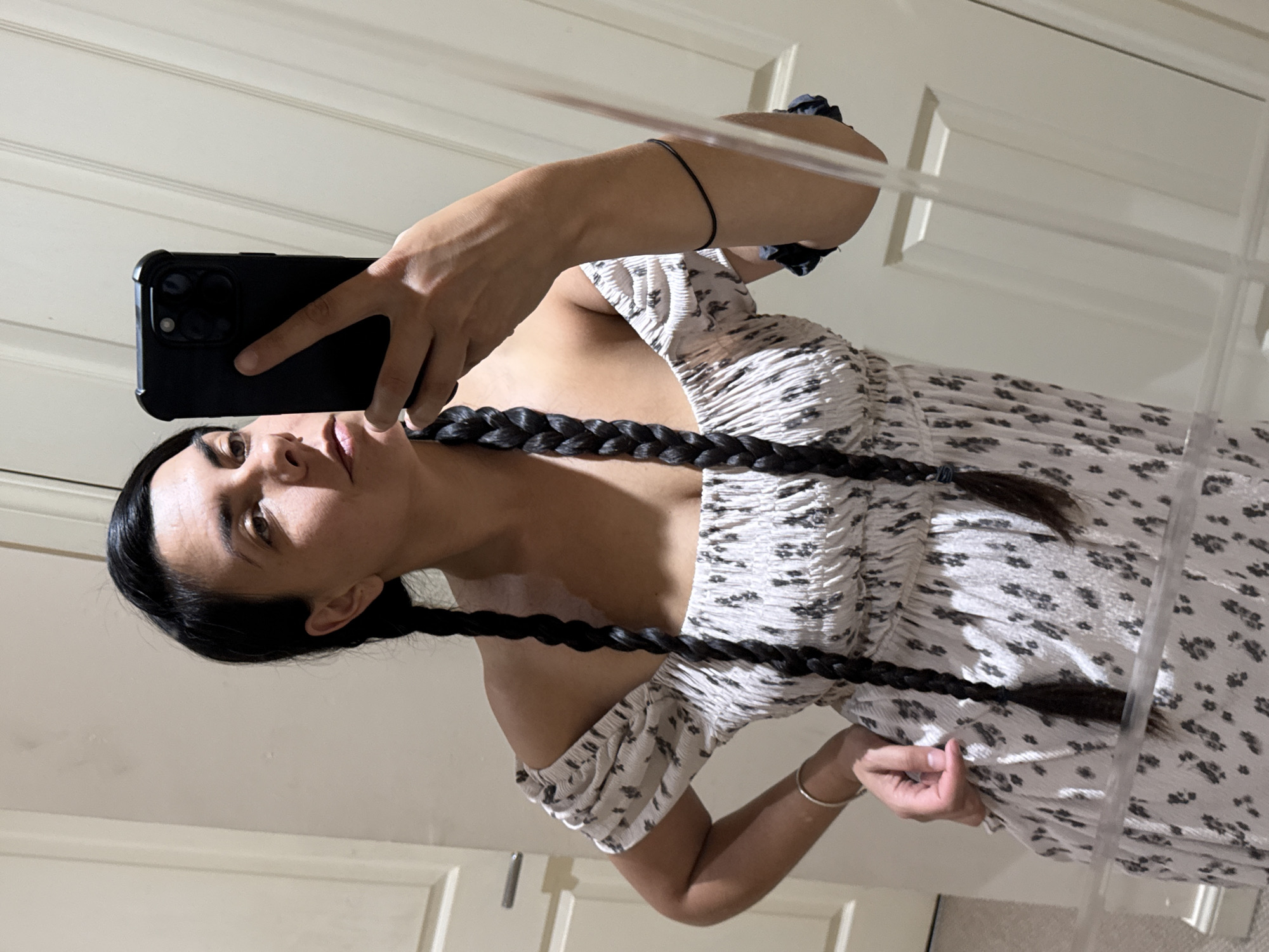 The image shows a woman taking a mirror selfie. She has dark hair styled in two braids and is wearing an off-the-shoulder dress with a floral pattern. She is holding a black smartphone in her right hand, capturing the reflection. The background appears to be a room with light-colored doors and walls, suggesting an indoor setting. There are no discernible landmarks or context that would allow for pinpointing a specific location within Great Yarmouth.