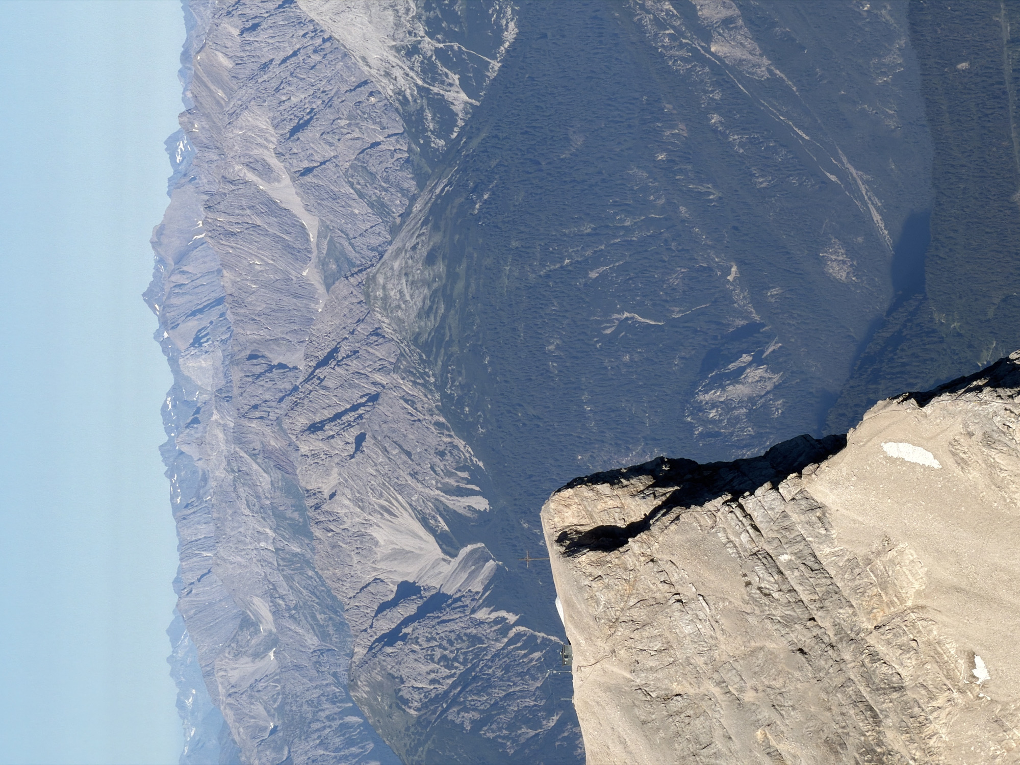 This image depicts a majestic mountain landscape with a sharp, rocky peak in the foreground. The peak is a light, sandy color and shows clear stratification, indicating geological layers. On the peak, there's a small, dark structure and a prominent cross. The background reveals a vast expanse of rugged, grey mountains stretching to the horizon under a clear, pale blue sky. The lower slopes of the mountains are covered in dense, dark green coniferous forests. The scene captures the grandeur of alpine scenery, likely from a high vantage point. This location is identifiable as the Zugspitze, Germany's highest peak, situated near the town of Grainau in the Bavarian Alps.