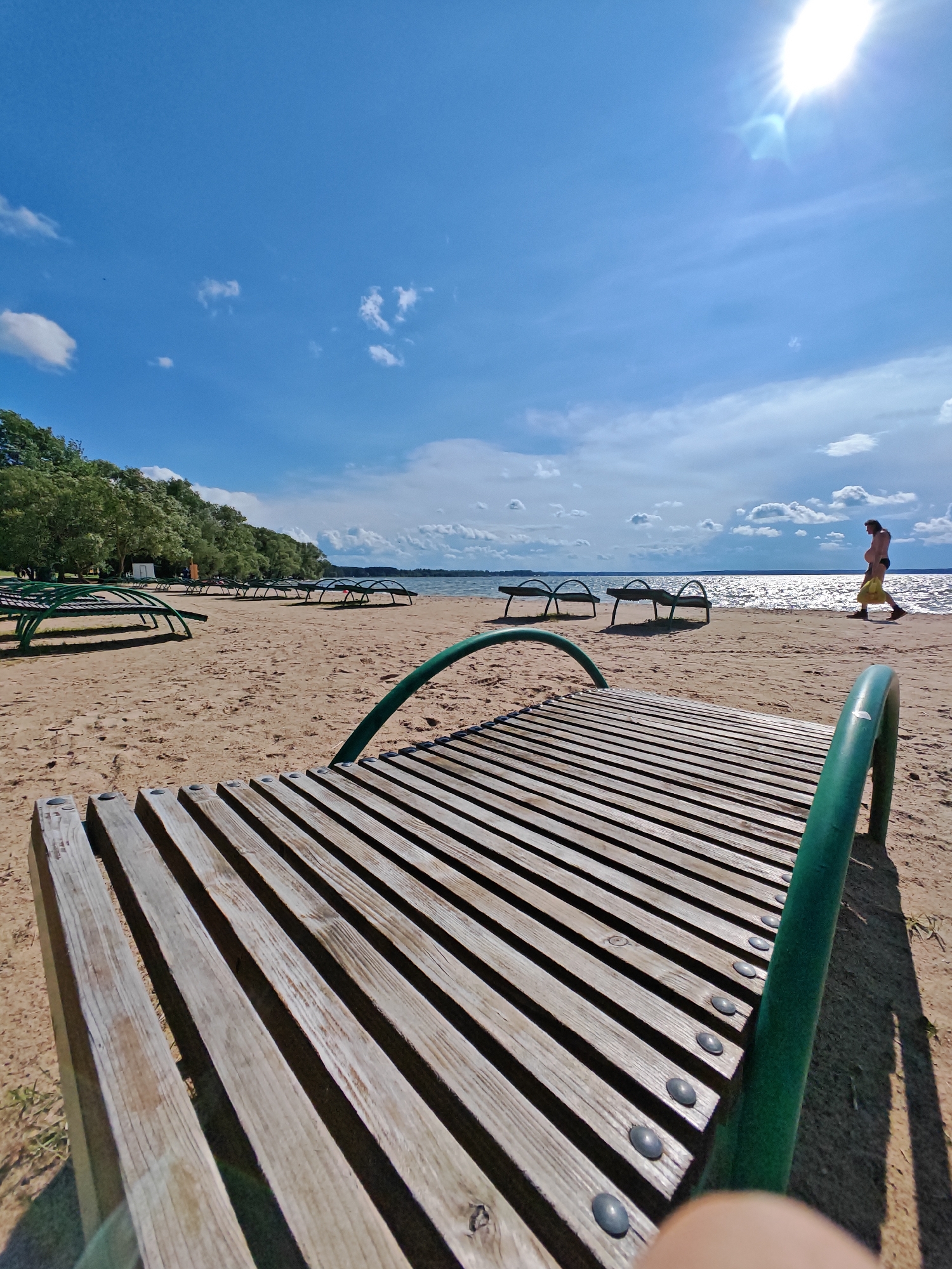 In the city of Ждановіцкі сельскі Савет, Belarus, a sun-drenched beach is depicted with numerous empty lounge chairs scattered across the sand. The foreground is dominated by a close-up view of a wooden lounge chair with green metal supports. In the middle ground, more of these chairs are visible, leading towards a body of water. A lone individual, wearing swimwear and carrying a yellow bag, walks along the shoreline in the background. The sky above is a vibrant blue, dotted with white clouds, and the sun is shining brightly, casting shadows on the sand. Trees line the far edge of the beach, and the water appears calm with sunlight reflecting off its surface.