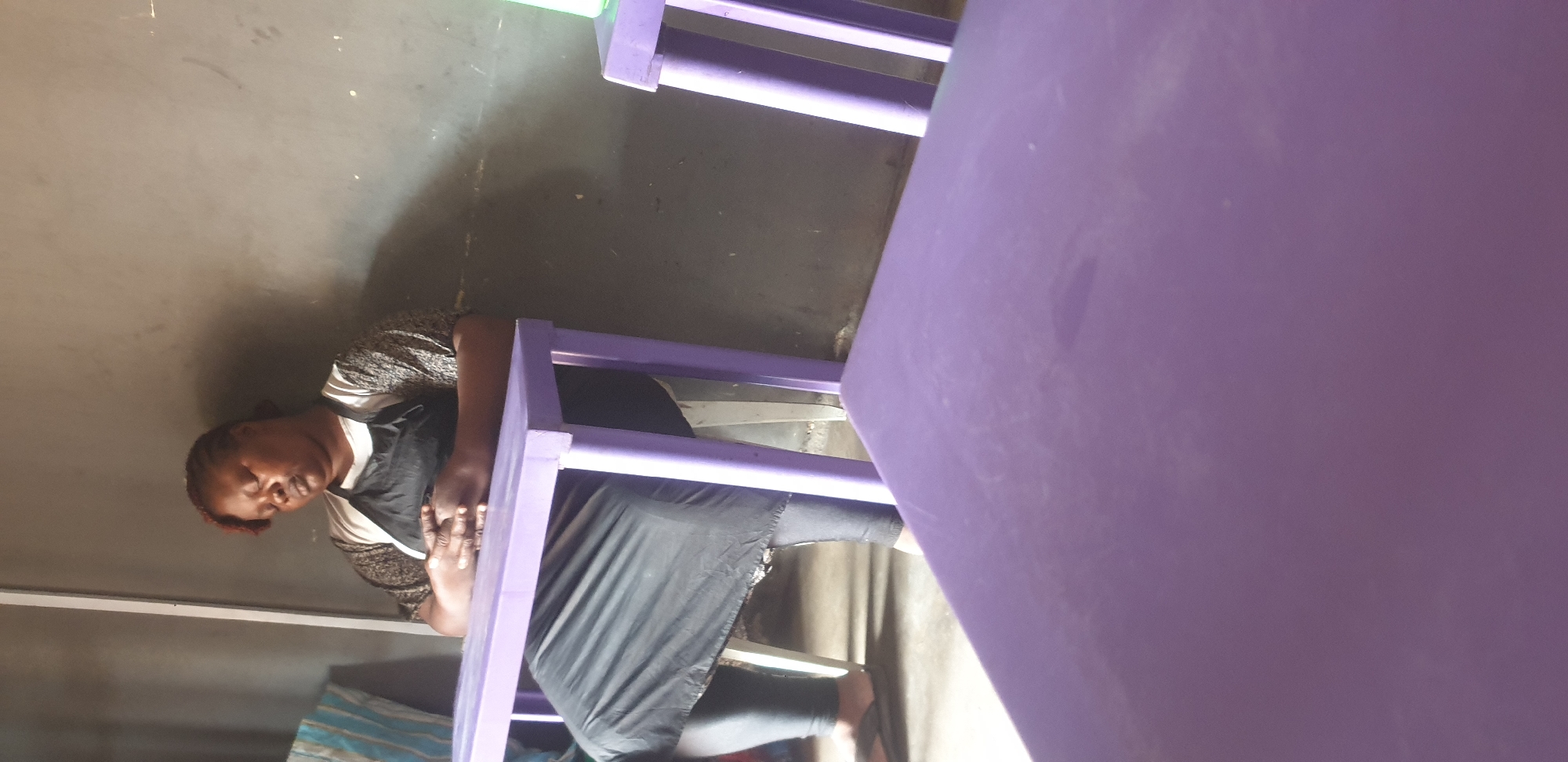 A woman with her eyes closed is seated at a purple table, wearing an apron over a white and patterned shirt. Her hands are clasped in her lap. She is wearing dark leggings and flip-flops. The background consists of a grey wall. The scene appears to be indoors, possibly a restaurant or cafe, with another purple table partially visible in the foreground. The overall lighting is dim.