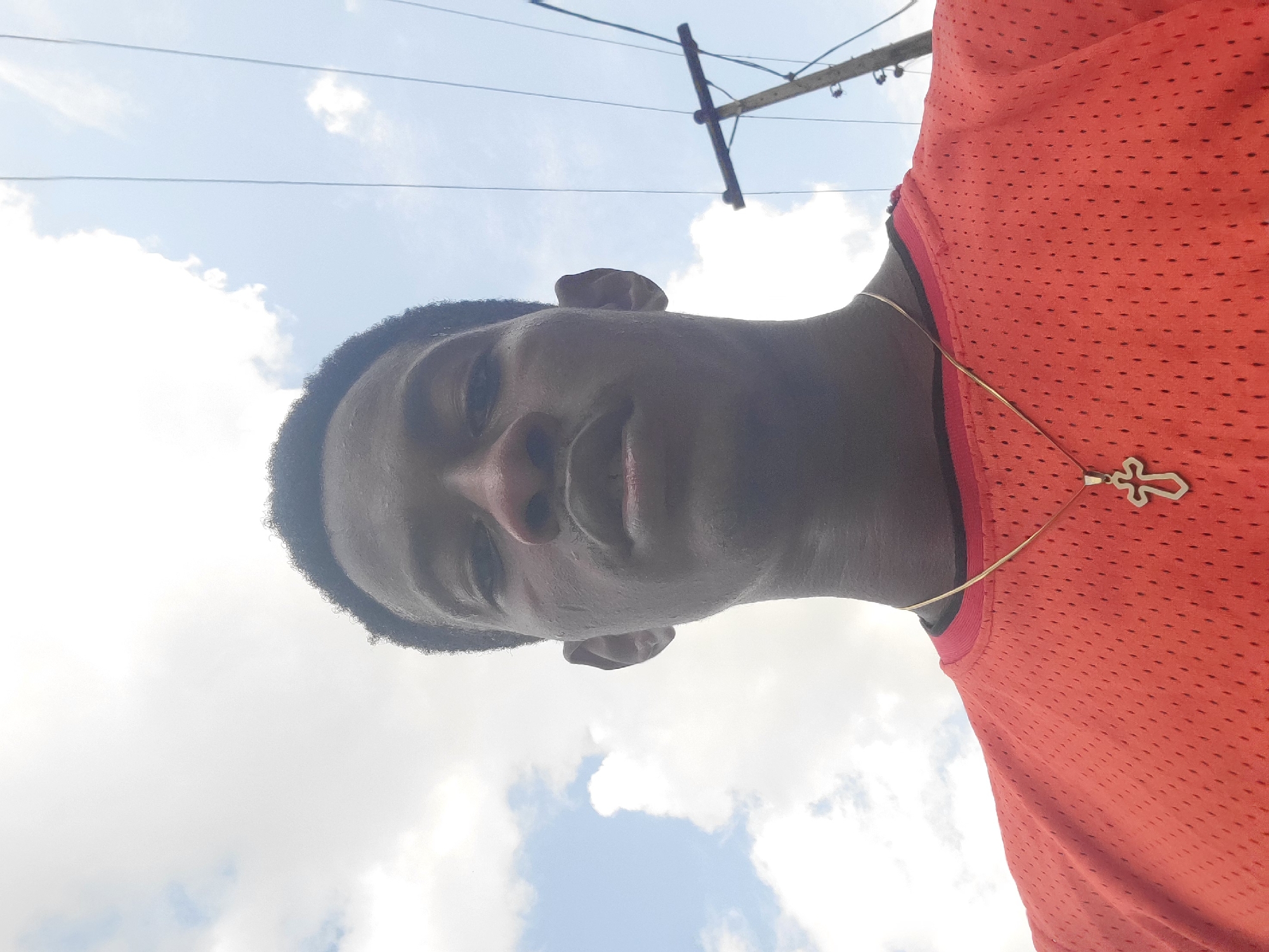 The image is a low-angle, close-up shot of a young Black male, taken outdoors against a bright, cloudy sky. The subject is wearing a bright orange mesh t-shirt and a gold chain with a cross pendant. His hair is short and dark, and he is looking directly at the camera with a relaxed expression.
The sky is mostly clear with scattered white clouds, suggesting it is daytime, likely midday due to the brightness. A utility pole with wires is visible in the background, indicating an urban or suburban setting. The image appears to be a selfie, given the perspective.
No text is visible in the image. The scene is casual and portrays a moment of personal documentation. The image has been rotated 90 degrees counter-clockwise.