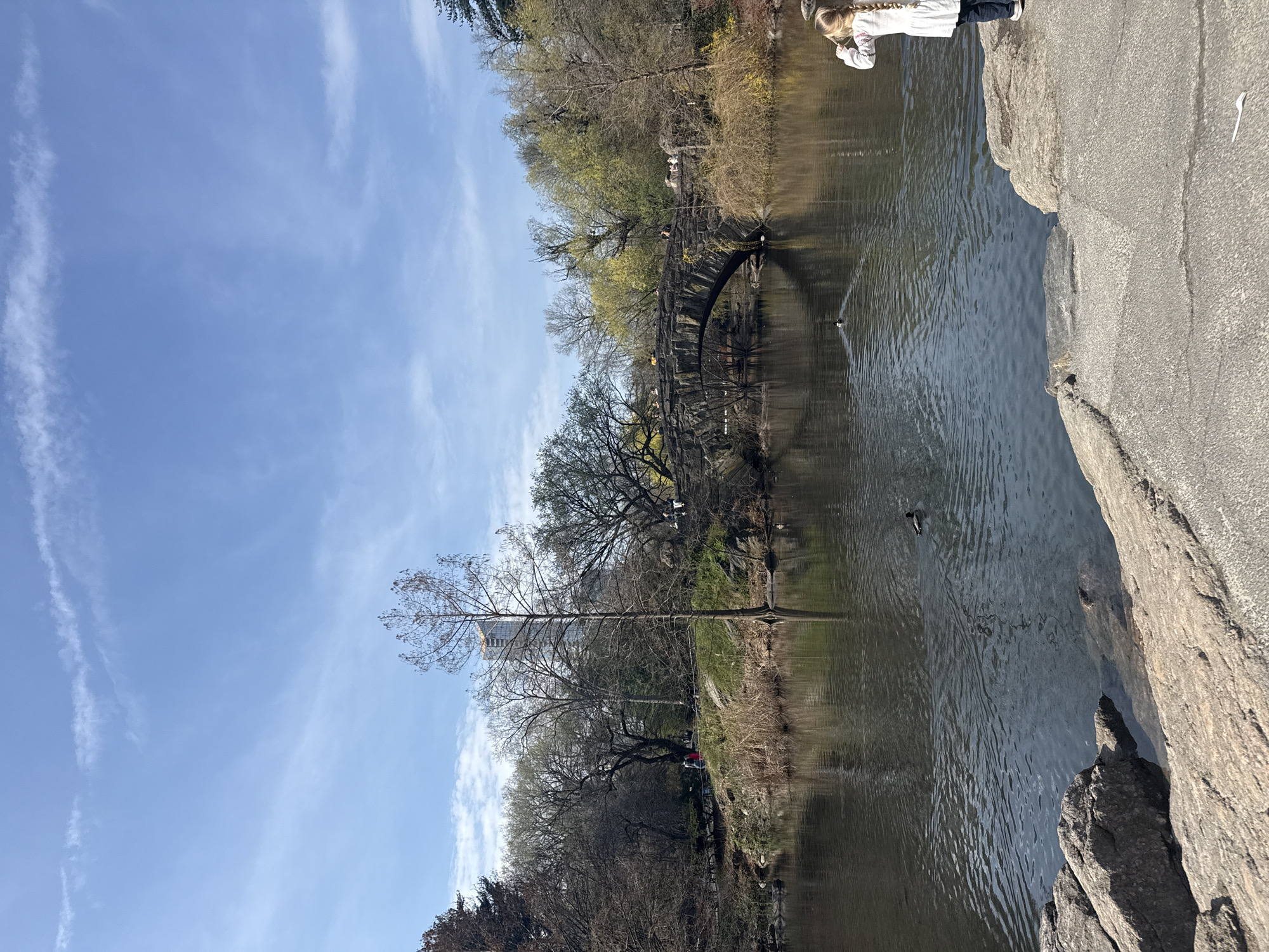 A horizontal outdoor shot depicts a body of water, a bridge, and landscape under a blue sky with wispy clouds. A young girl is standing on the rocks in the upper right corner. The bridge is arched and appears to be made of stone or a similar material. Trees line the edge of the water, some with full foliage and others still bare. The image is likely taken in Central Park, New York City.
