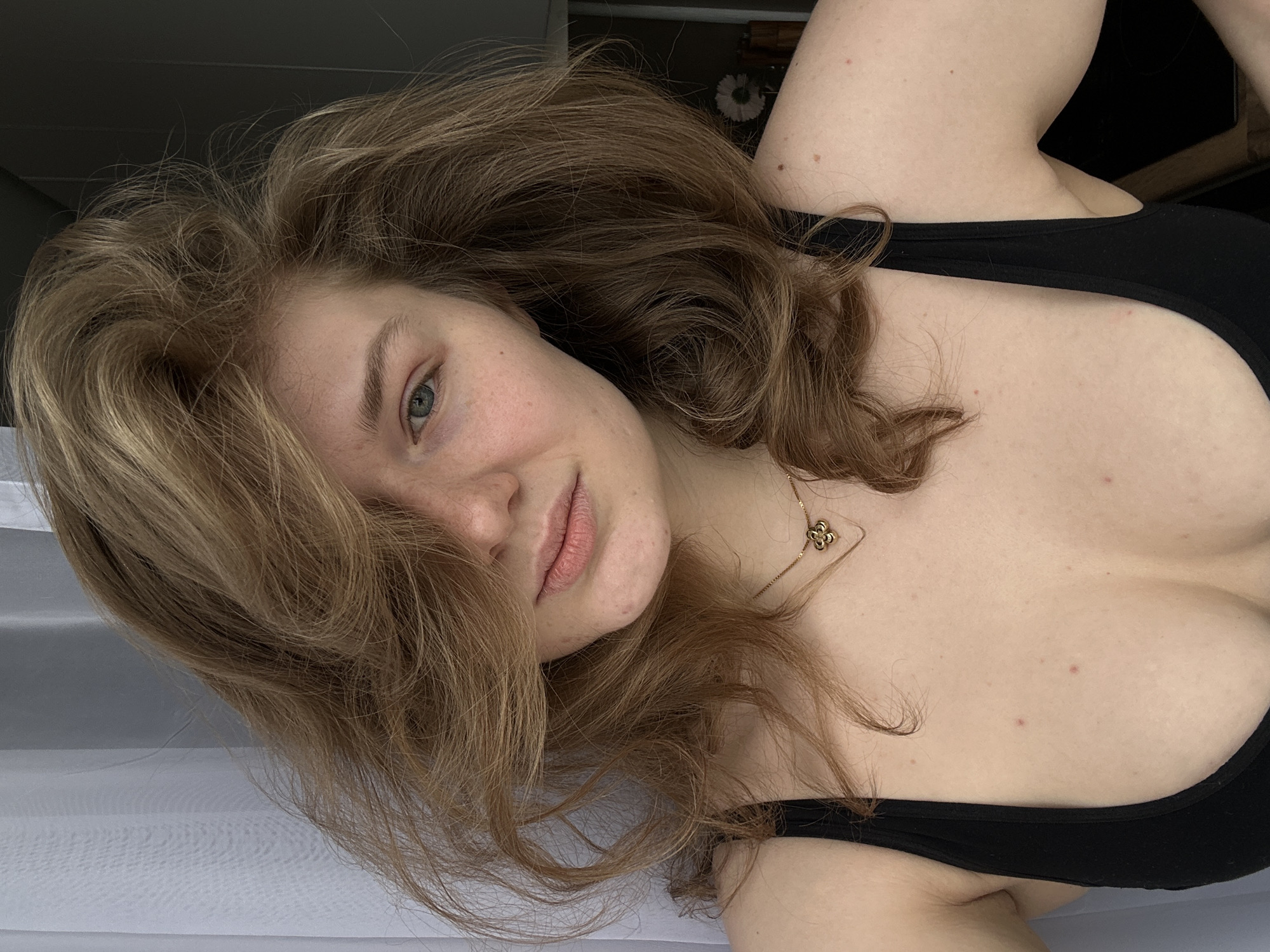 A woman with fair skin and wavy brown hair is lying down, gazing into the camera. She is wearing a black low-cut top and a delicate gold necklace. The background appears to be a lightly colored or white surface. There's no evidence to suggest the image was taken in Kazan, Russia.