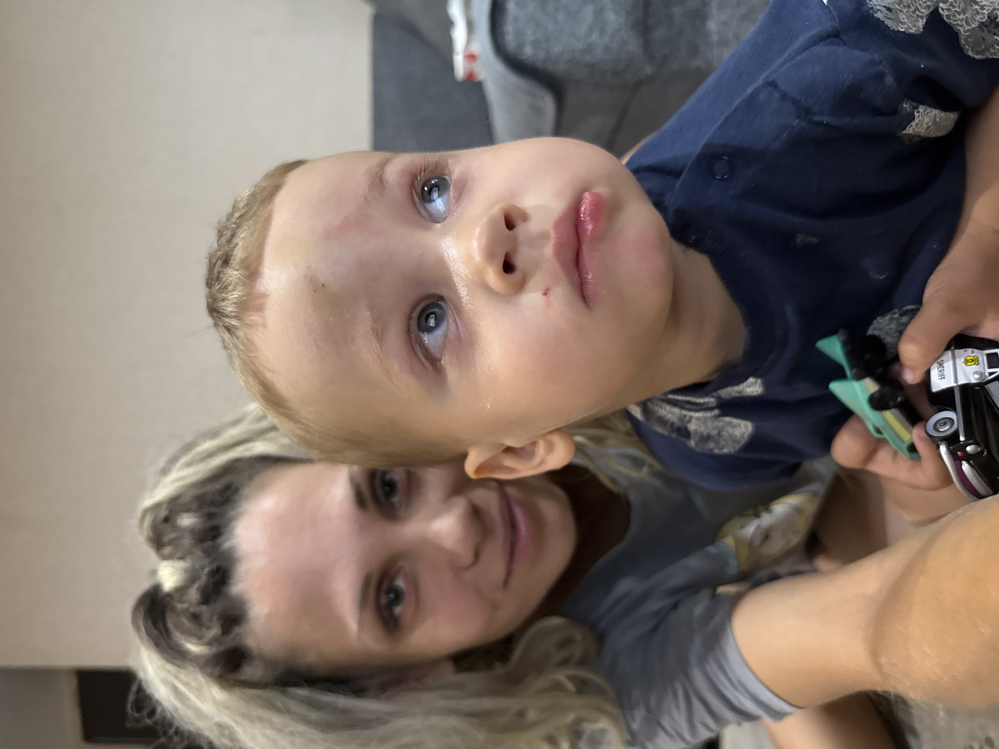 In Rusanovo, Russia, a close-up image captures a woman and a young child. The woman, positioned on the left, has blonde hair pulled back and is looking towards the camera with a slight smile. She is wearing a grey t-shirt. The child, on the right, is facing away from the camera, looking upwards and to the right. The child has blond hair and appears to have a minor injury on his forehead and a mark on his lower lip. He is wearing a dark blue shirt. The child's right hand is holding two toy cars. The background is blurred, showing a grey sofa and a light-colored wall. The lighting suggests an indoor environment, possibly a home.