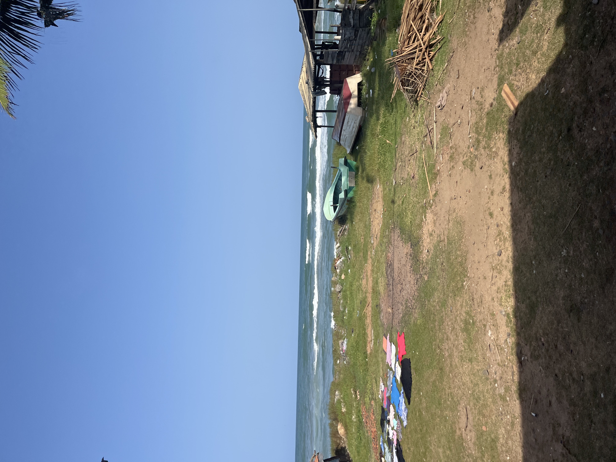 The image depicts a coastal scene in Moratuwa, Sri Lanka. The foreground shows a patch of dry, grassy ground with some scattered debris and a pile of wooden sticks. To the left, a collection of brightly colored laundry is laid out on the grass to dry.
In the middle ground, a small embankment separates the land from the ocean. Two small boats are visible on the grass near the embankment. One is green and upright, while the other is inverted and appears to be white with a red trim.
Further to the right, there is a rustic, open-sided structure with a corrugated metal roof, suggesting a small shelter or vendor stall. The sea stretches out to the horizon under a clear blue sky. Gentle waves are breaking on the shore, creating a serene coastal atmosphere. The image is taken during daylight hours, likely mid-morning or afternoon, given the angle of the sunlight casting shadows. There are no people visible in the frame.