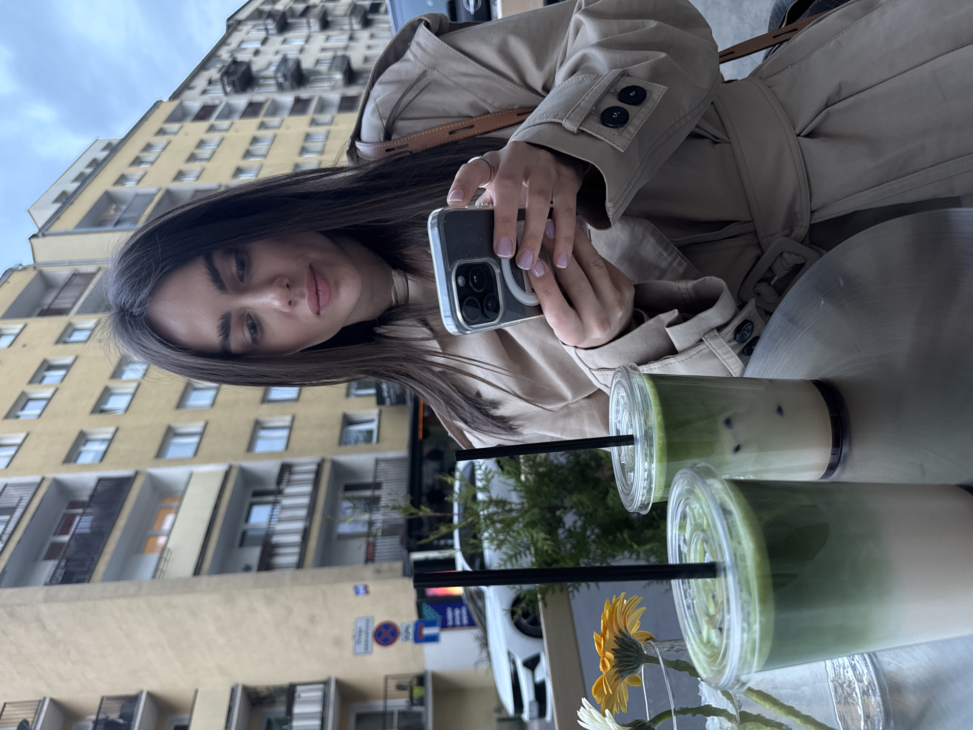 A woman in a beige trench coat holds a smartphone in her hands, poised as if taking a picture or video. She is seated at an outdoor table with two cups of green matcha lattes in front of her, along with a small vase of yellow and white gerbera daisies. A tall, pale yellow apartment building typical of Warsaw architecture is visible in the background. Street signs and cars suggest the location is in an urban area of Warsaw, Poland.