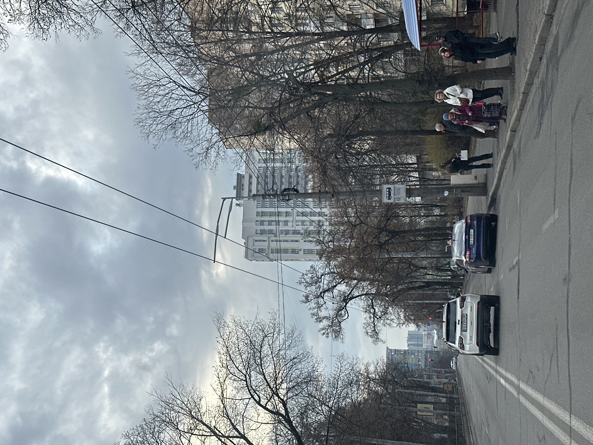 The image depicts a street scene in Kyiv, Ukraine, on an overcast day. The street is paved and has double white lines running down the center. Two cars are visible in the foreground: a white car with license plate number K023CO2 is in the lower half of the image and a dark blue car is behind it. Bare trees line both sides of the street, obscuring some of the buildings. A tall, white building is visible behind the trees and in the distance other buildings can be seen. On the right sidewalk, several people are walking. There is also a bus stop. Overhead power lines are visible against the gray sky.