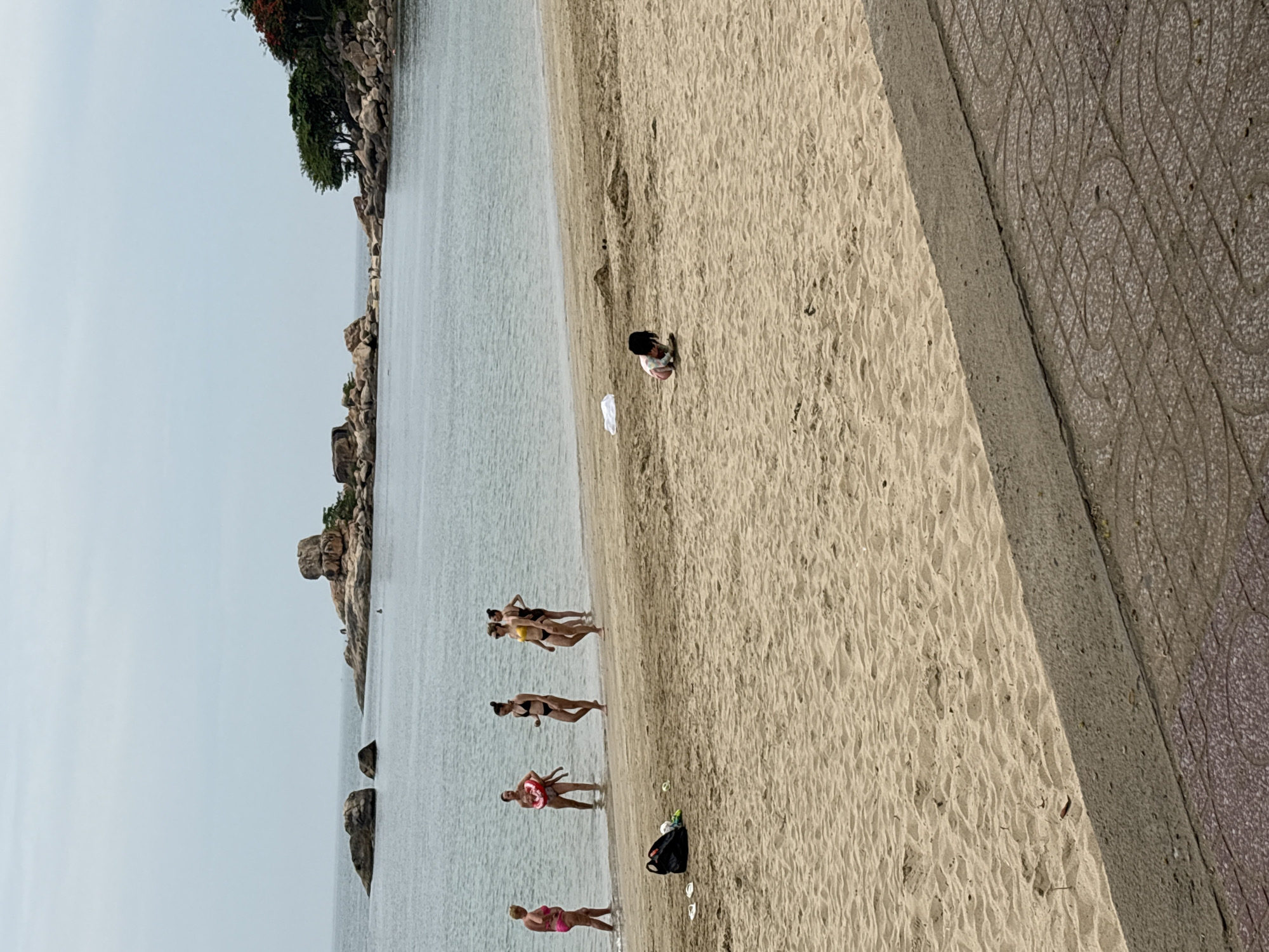 The image depicts a beach scene in Bac Nha Trang Ward, Vietnam. The view is a high-angle shot, likely taken from an elevated position along a paved promenade. The scene includes the beach, the water, and a rocky coastline in the distance.
Several people are visible along the shoreline: a small group of women are standing in the water, while another individual is crouching on the beach. A black bag and some other items are on the sand near the water's edge. The beach itself is sandy, showing footprints and other impressions. The water appears calm and clear. On the left of the frame, there is a rocky area with greenery. The right edge of the frame shows a paved area with a patterned surface.