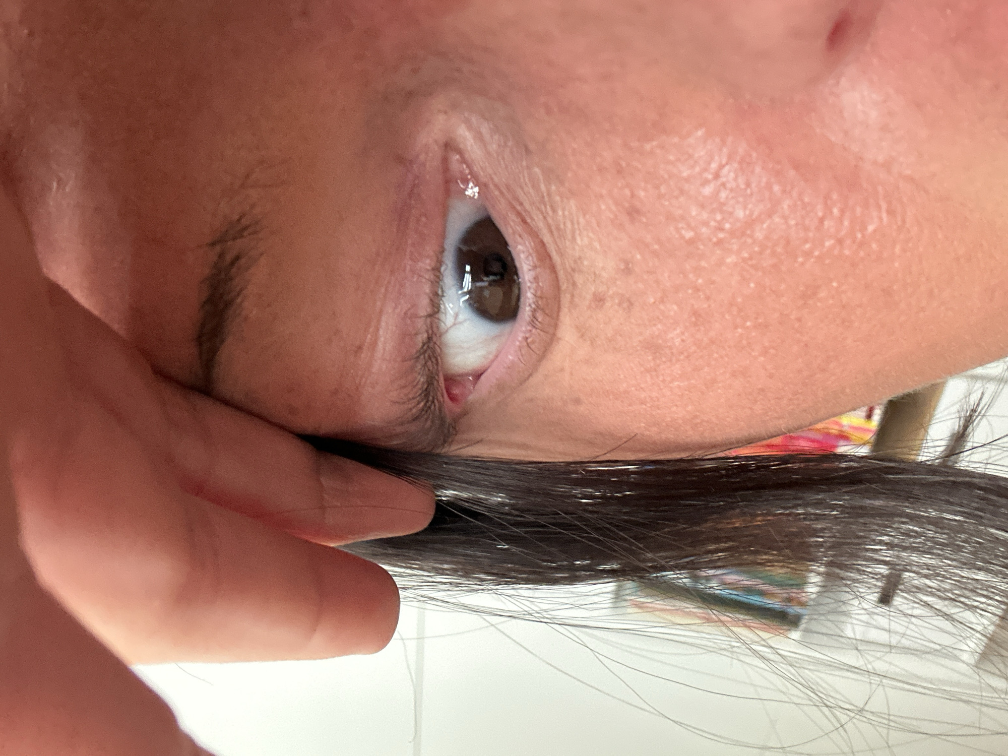 The image shows a close-up of a person's eye. The eye is brown, and the person's skin is light-skinned. Some hair strands are visible in the lower portion of the frame. A finger is holding the hair up. There is no information in the image to relate it to the city of Wealden, United Kingdom.