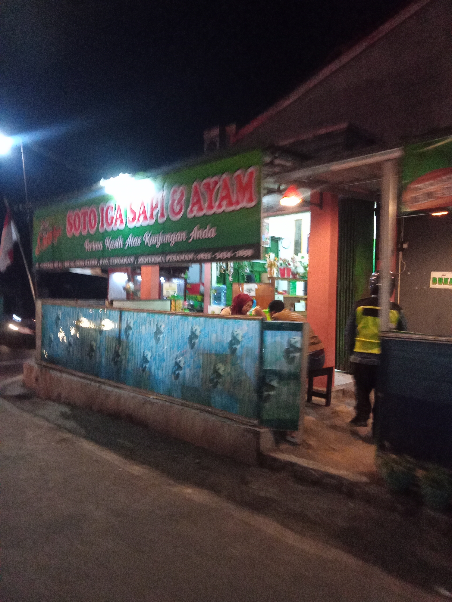 The image depicts the exterior of a food establishment, "SOTO ICA SAPI & AYAM," located in Tengaran, Indonesia. The sign is a prominent green banner with red and white lettering. Below the main sign, a smaller banner states, "Terima Kasih Atas Kunjungan Anda" (Thank you for your visit) and provides contact information: "MENERIMA PESANAN: 0821-3434-1859". The establishment appears to be a simple building with an open front area, suggesting a casual dining setting.
In the foreground, a blue barrier with a repeating pattern of pandas and bamboo is visible. Behind this barrier, two individuals are seated at tables. One person, wearing a red headscarf, is looking down, possibly eating. The other person, partially visible, is facing away from the camera.
Behind the seated patrons, an orange-painted pillar supports a portion of the building's overhang. Further back, a person wearing a yellow and black safety vest is standing near an open doorway, appearing to be an employee or attendant. The doorway leads into a dimly lit interior where shelves stocked with various items are visible, along with a red light fixture hanging from the ceiling.
The environment is dark, indicating it is nighttime, with artificial lighting illuminating the scene. A street or pathway is visible in the lower portion of the image, and a faint shadow is cast across it. The overall impression is of a modest, local eatery operating in the evening.