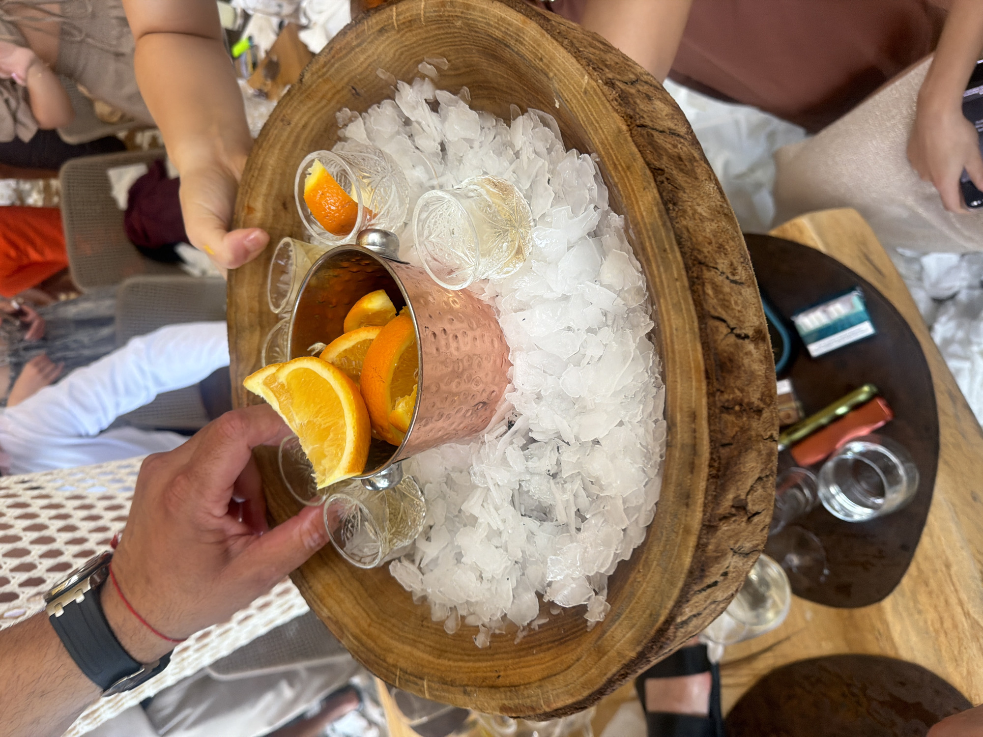 A close-up, overhead shot captures a wooden platter filled with crushed ice, serving as a bed for several small glasses of clear liquid. The glasses are adorned with orange slices, some whole and some cut into smaller pieces. A copper-colored metal cup, also containing orange slices, sits in the center of the platter. The platter is held by two individuals, one's hand is visible on the left side, wearing a watch and a red string bracelet. The other individual's hand is on the right side, gently holding the edge of the platter. The background is blurred but suggests an outdoor or semi-outdoor setting with glimpses of people and furniture.
