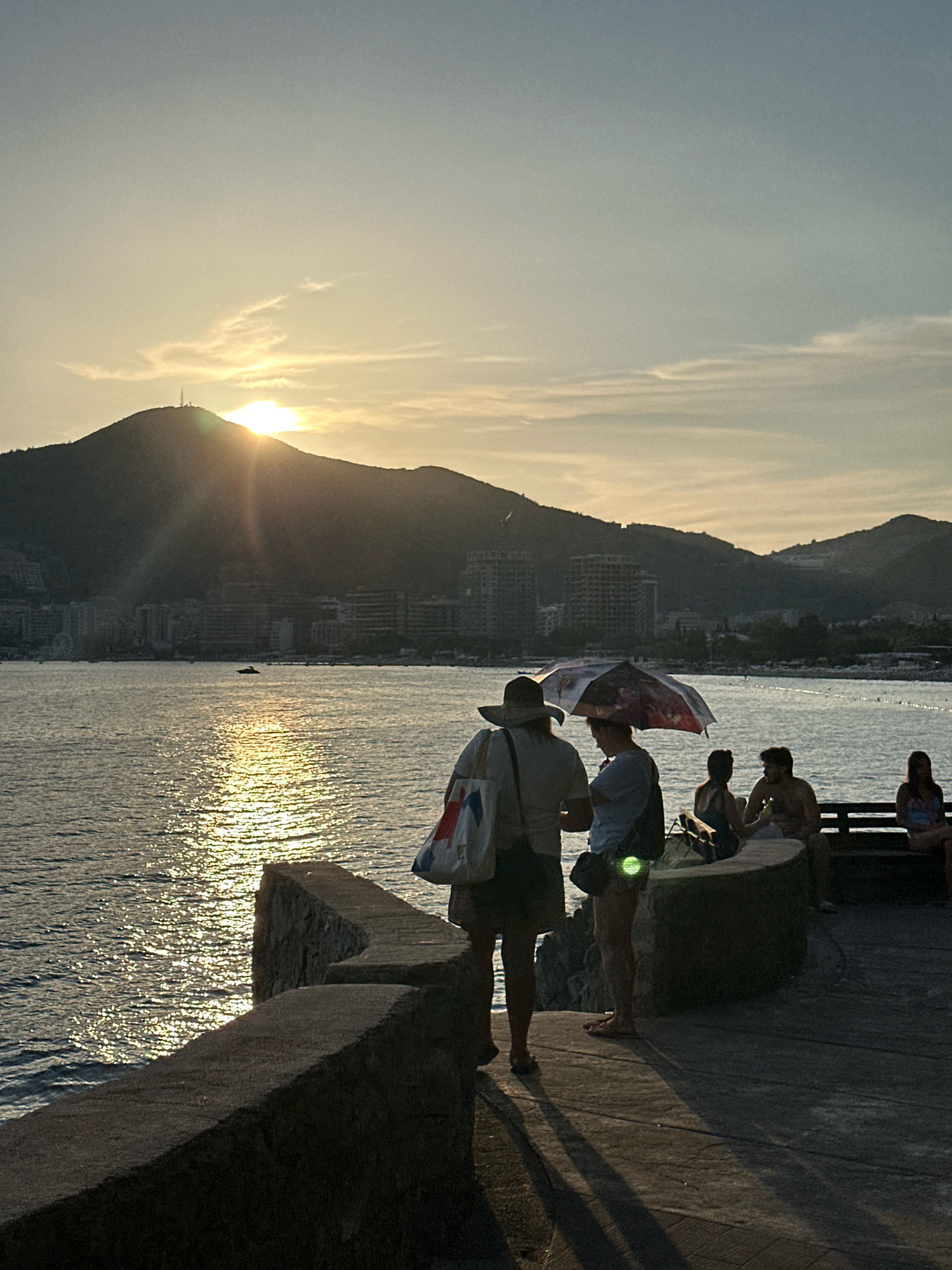 The image depicts a sunset scene in Budva, Montenegro. The sun is low on the horizon, casting a warm glow across the sky and reflecting on the calm sea. A prominent mountain dominates the background, with the silhouette of a city visible at its base. In the foreground, several individuals are gathered on a paved walkway near a stone wall overlooking the water. Two people stand under an open umbrella, engaged in conversation, while others are seated on a bench. The lighting suggests it is late afternoon or early evening, with long shadows stretching across the walkway. The overall atmosphere is serene and tranquil, capturing a moment of relaxation by the coast.