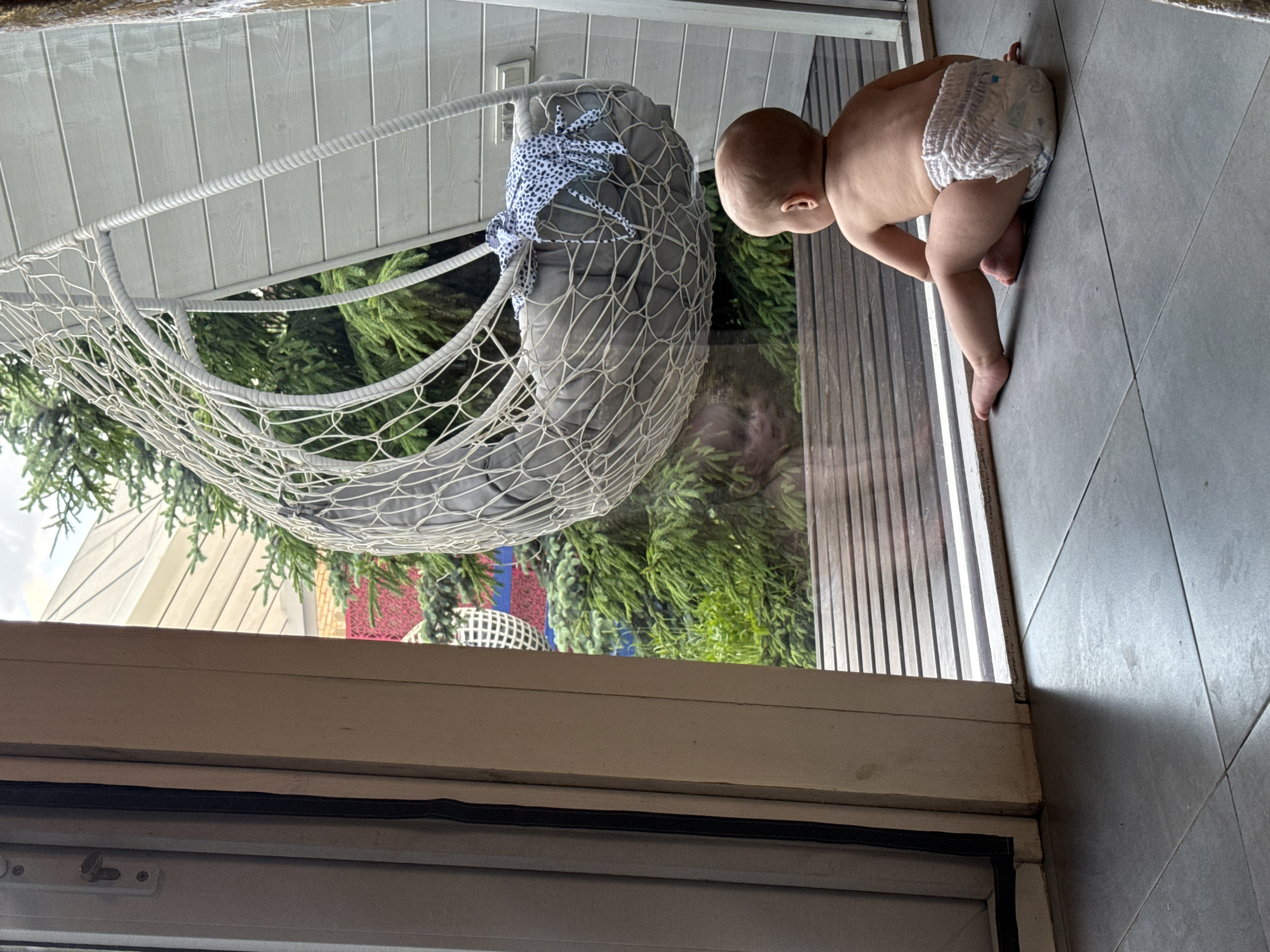 A baby in a diaper is crawling on a gray tiled floor in what appears to be an indoor setting next to a sliding glass door. Outside the door, there's a hanging white woven chair with a gray cushion and blue polka-dot ties, along with trees and parts of a building. The bottom of the door frame is visible in the foreground. The location is unknown.