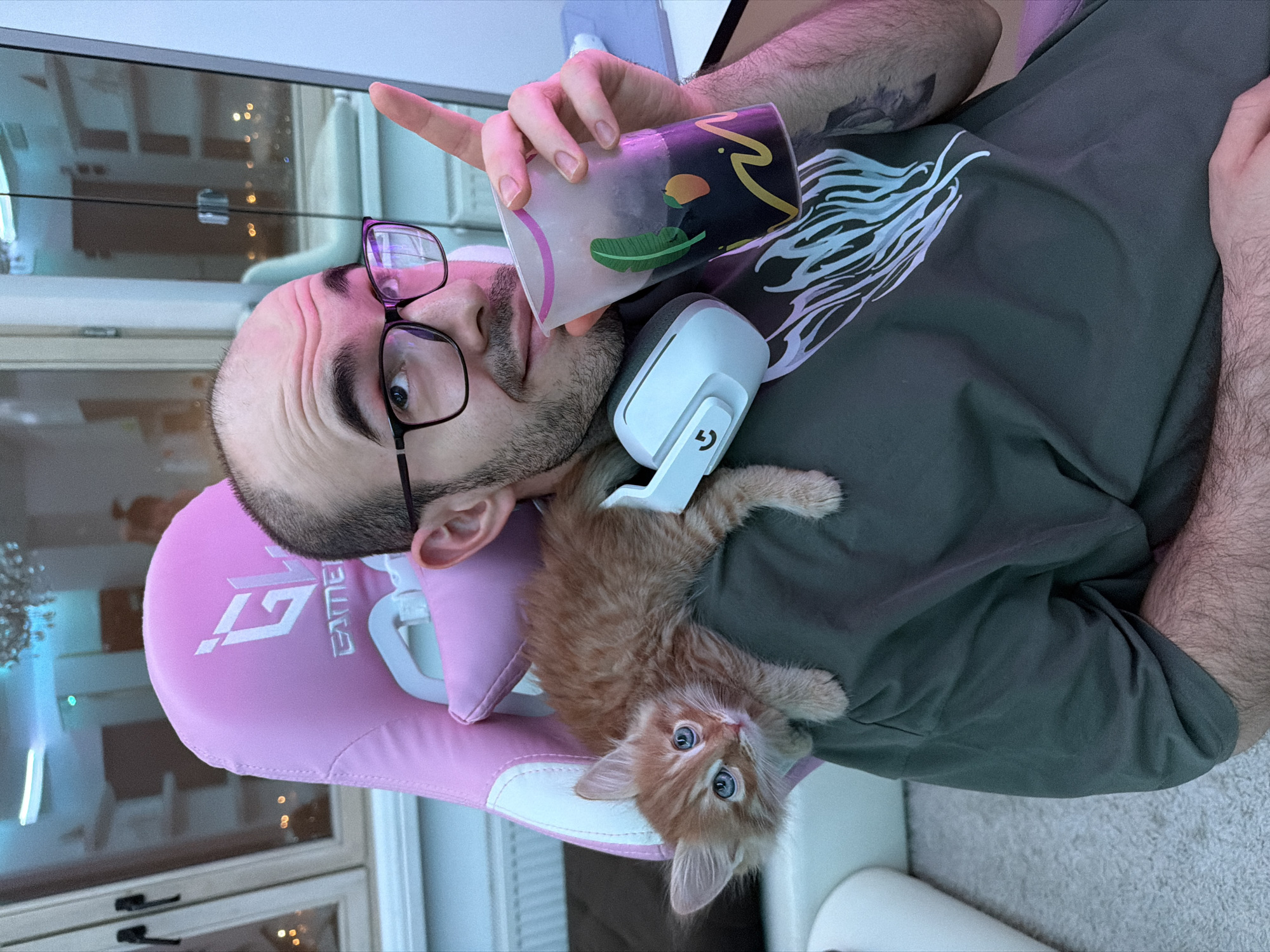 This image, set in Khimki, Russia, captures a man and a kitten in a relaxed indoor setting. The man, with a trimmed beard and dark-framed glasses, is seated in a vibrant pink gaming chair. He wears a dark olive-green t-shirt featuring a stylized light blue and white graphic design on the chest. Around his neck is a white Logitech G headset. He holds a clear cup with a black abstract pattern, adorned with a green leaf and an orange fruit design, and is pointing his index finger upwards. His expression is one of mild surprise or curiosity as he looks directly towards the viewer.
Resting on the man's shoulder and chest is a fluffy orange kitten with captivating blue eyes. The kitten is also looking towards the camera with a curious gaze. The pink gaming chair has white accents and clearly displays "Gamer" text with a prominent "G" logo.
The background reveals a room with a large window or reflective surface behind the man, through which blurred lights and subtle architectural details are visible, suggesting an exterior view or reflections from inside. A light-colored carpet is partially visible on the floor. The overall lighting has a cool, slightly blueish hue, hinting at artificial light sources or an evening shot. The scene conveys a warm, affectionate interaction between the man and his pet.