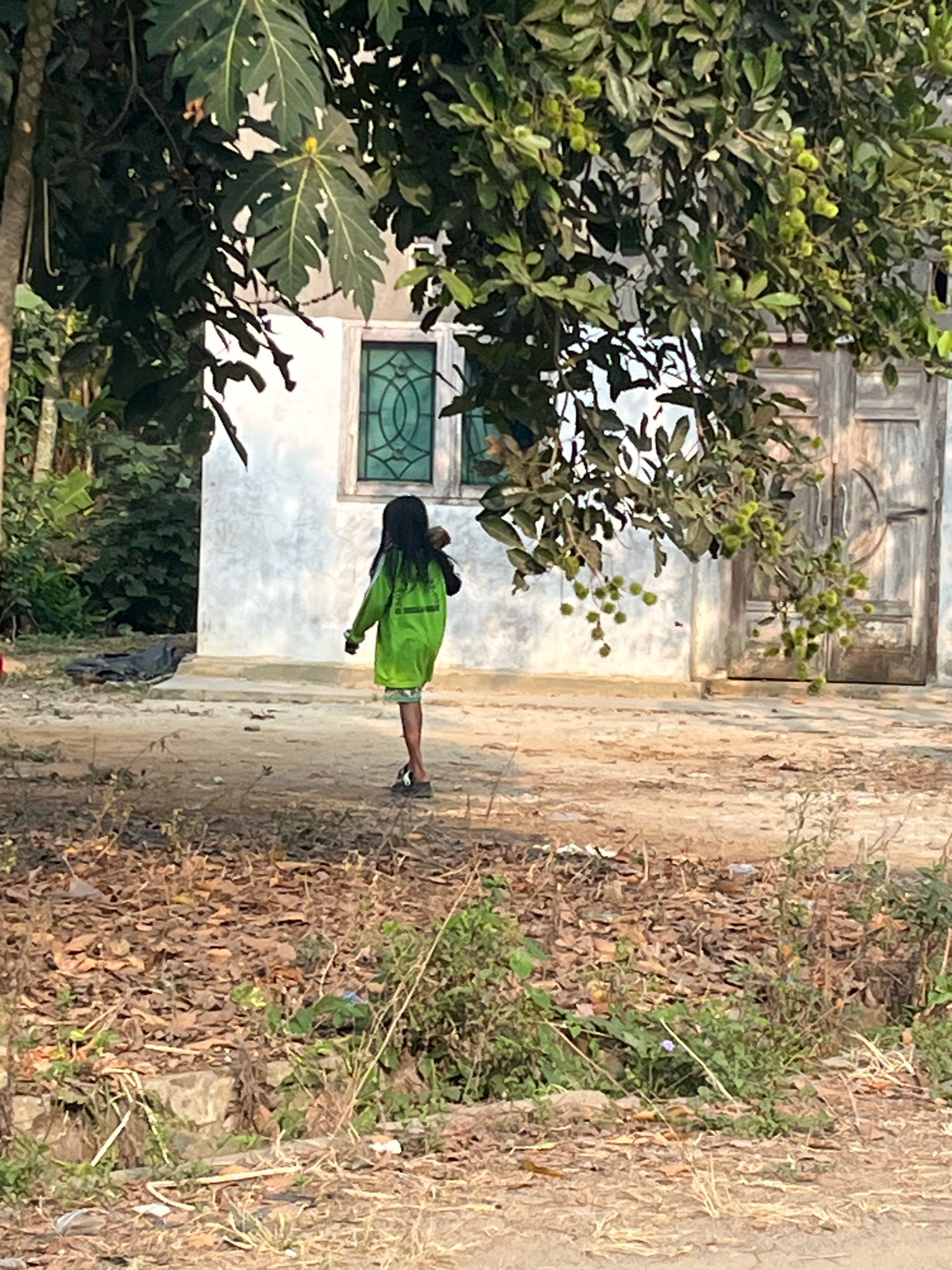 A young girl with long dark hair walks away from the camera, carrying a dark object over her left shoulder, in front of a white building with a green-framed window and a wooden door. She is wearing a bright green long-sleeved jacket over patterned shorts and dark sandals. The building appears to be in a rural setting, with trees and foliage surrounding it. The ground in the foreground is covered with dry leaves and sparse vegetation. The overall lighting suggests it is daytime. The specific location is in Margodadi, Indonesia.