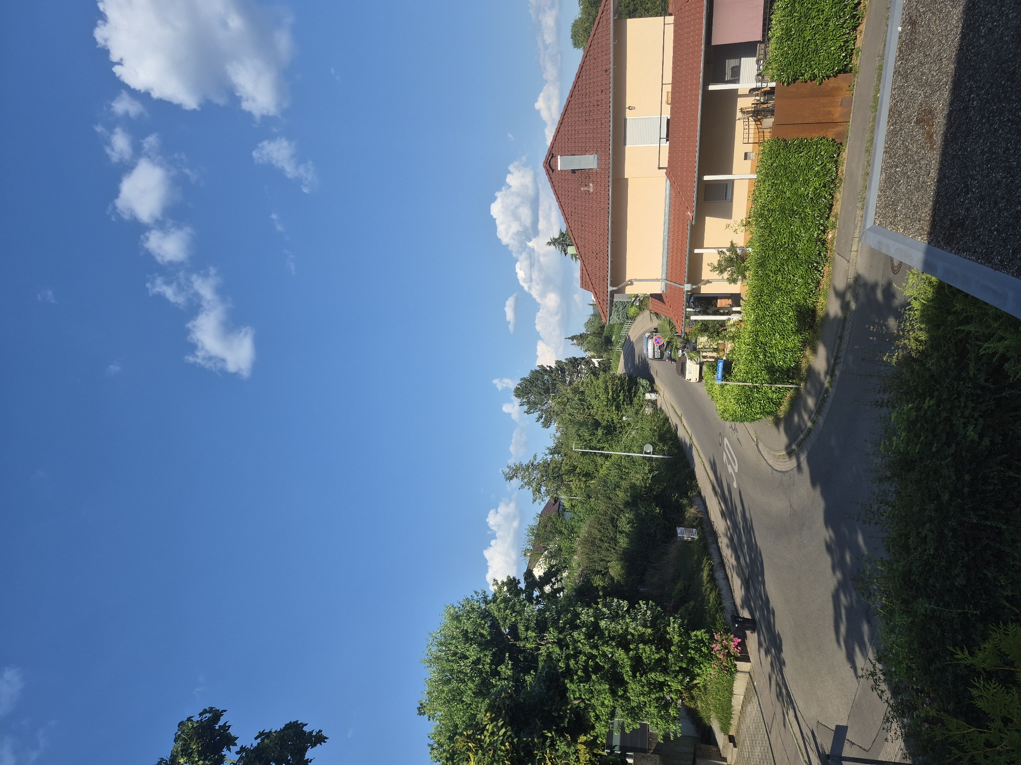 The image shows a residential street with houses and trees under a blue sky with white clouds. A paved road curves through the scene, with a speed limit sign indicating 30. On the right side of the road, a yellow house with a red tiled roof is visible, surrounded by a neatly trimmed green hedge. There are two cars parked on the road. On the left side of the road, a large green tree stands prominently. The environment appears to be a suburban or semi-rural area on a sunny day.