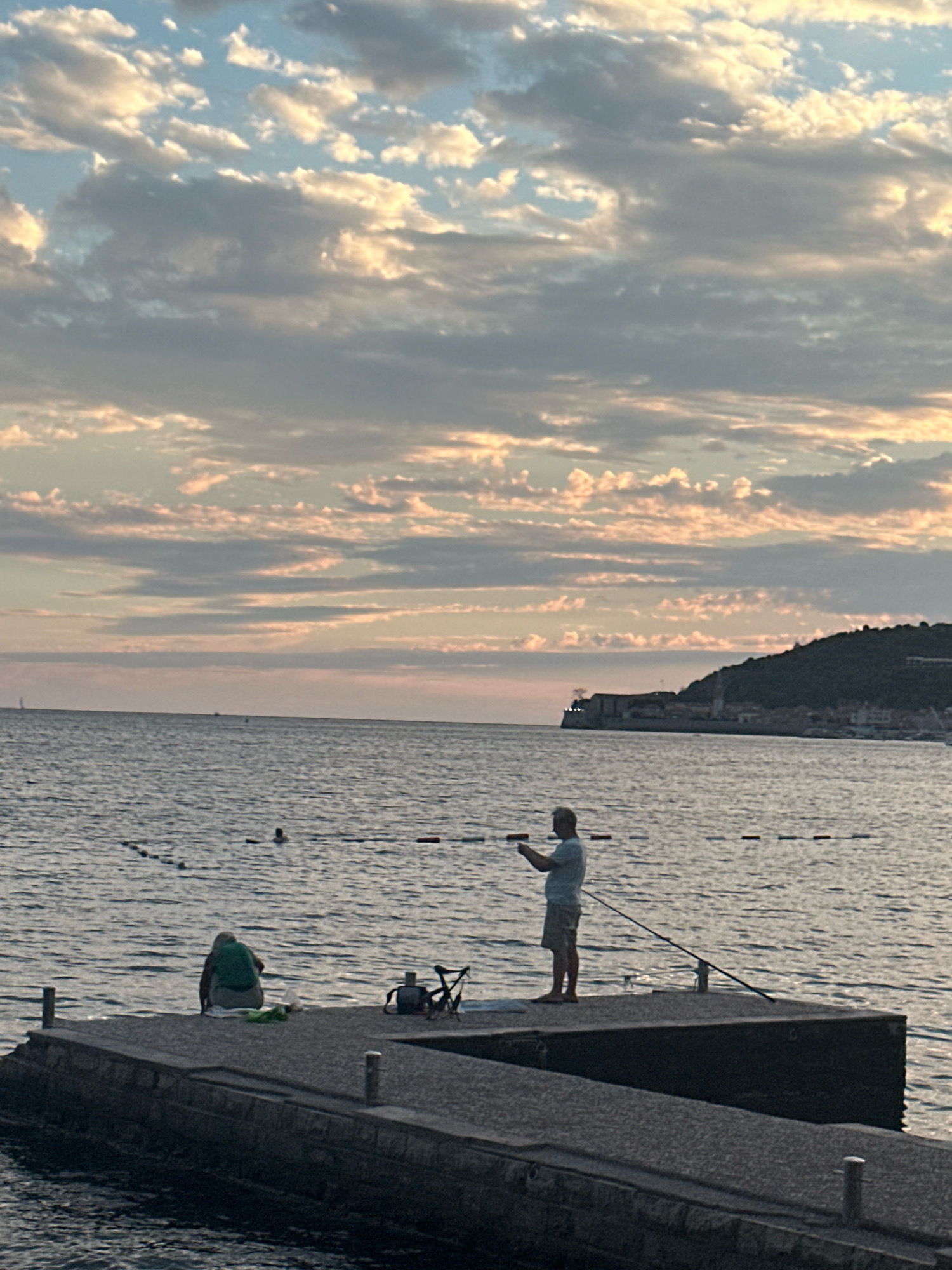 The image captures a serene evening scene on a pier in Budva, Montenegro. The sky is adorned with a tapestry of clouds, tinged with hues of pink and orange, suggesting either sunrise or sunset. The calm sea stretches out to the horizon, where a hillside town, characteristic of Budva's coastal landscape, is visible in the distance.
On the pier, two individuals are present. One person, seen from behind, is seated on the pier, wearing a green top and what appears to be a light-colored towel or blanket beside them. To their right, a bicycle and a bag are placed on the pier. The second person is standing on the edge of the pier, facing the water and holding a fishing rod, engaged in fishing. Their attire consists of a light-colored t-shirt and shorts. In the water, a few buoys mark a swimming area, and at least one swimmer is visible. The pier itself is made of concrete, with stone masonry visible on its sides. The overall atmosphere is peaceful, with the gentle lapping of waves and the soft light of the sky creating a tranquil ambiance.