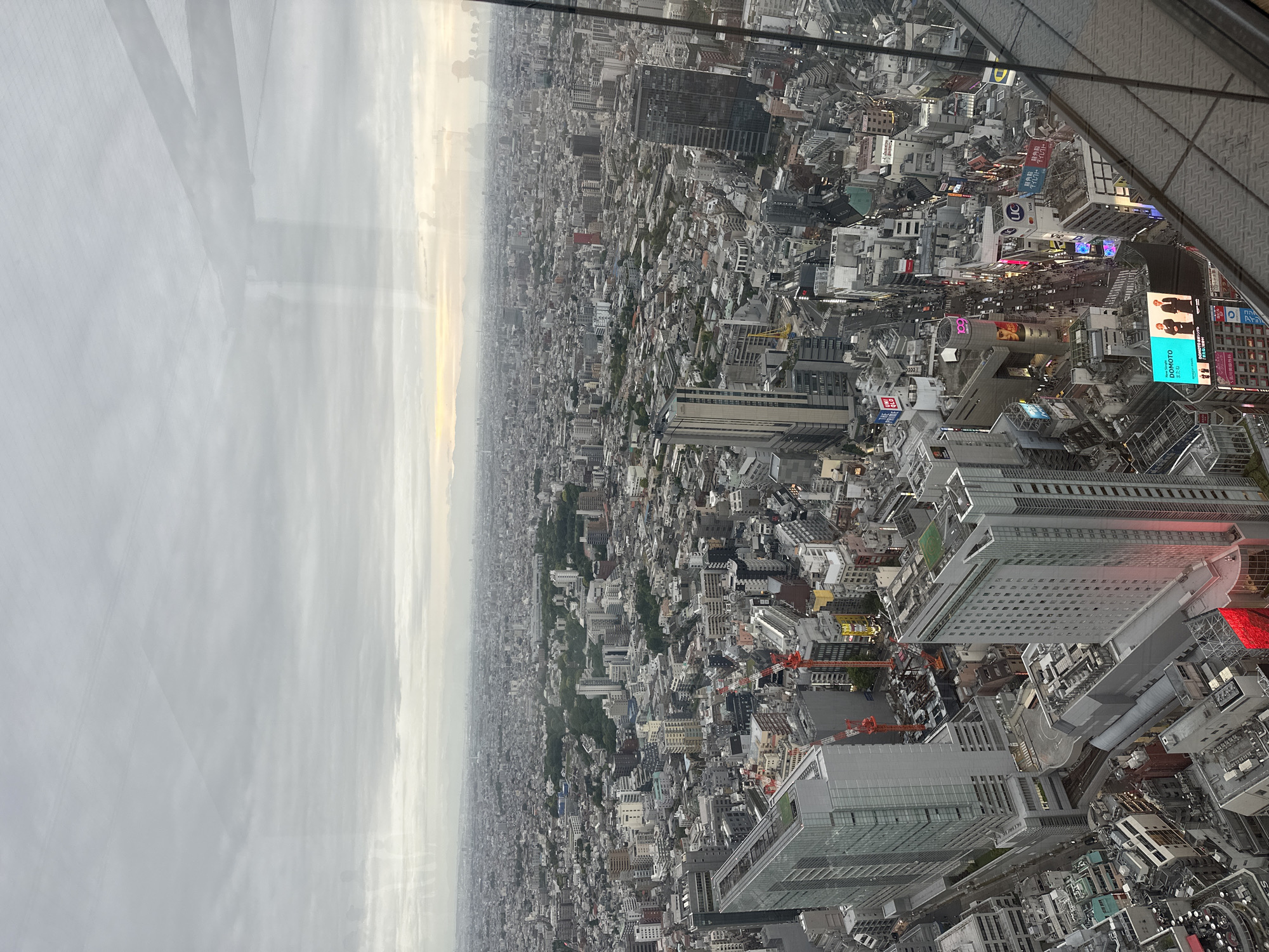 The image is an aerial view of Shibuya, Japan, from what appears to be a tall building. The sky is overcast and hazy. The dense urban landscape extends to the horizon with a high concentration of buildings varying in height, shape, and color. Near the bottom right corner, there are neon signs and billboards, indicating a commercial district. A building structure with a metal support system is visible in the upper right corner, suggesting the photograph was taken from within a structure. There are several construction cranes interspersed among the buildings, indicating ongoing development. In the distance, the city fades into a hazy skyline. The image is rotated.