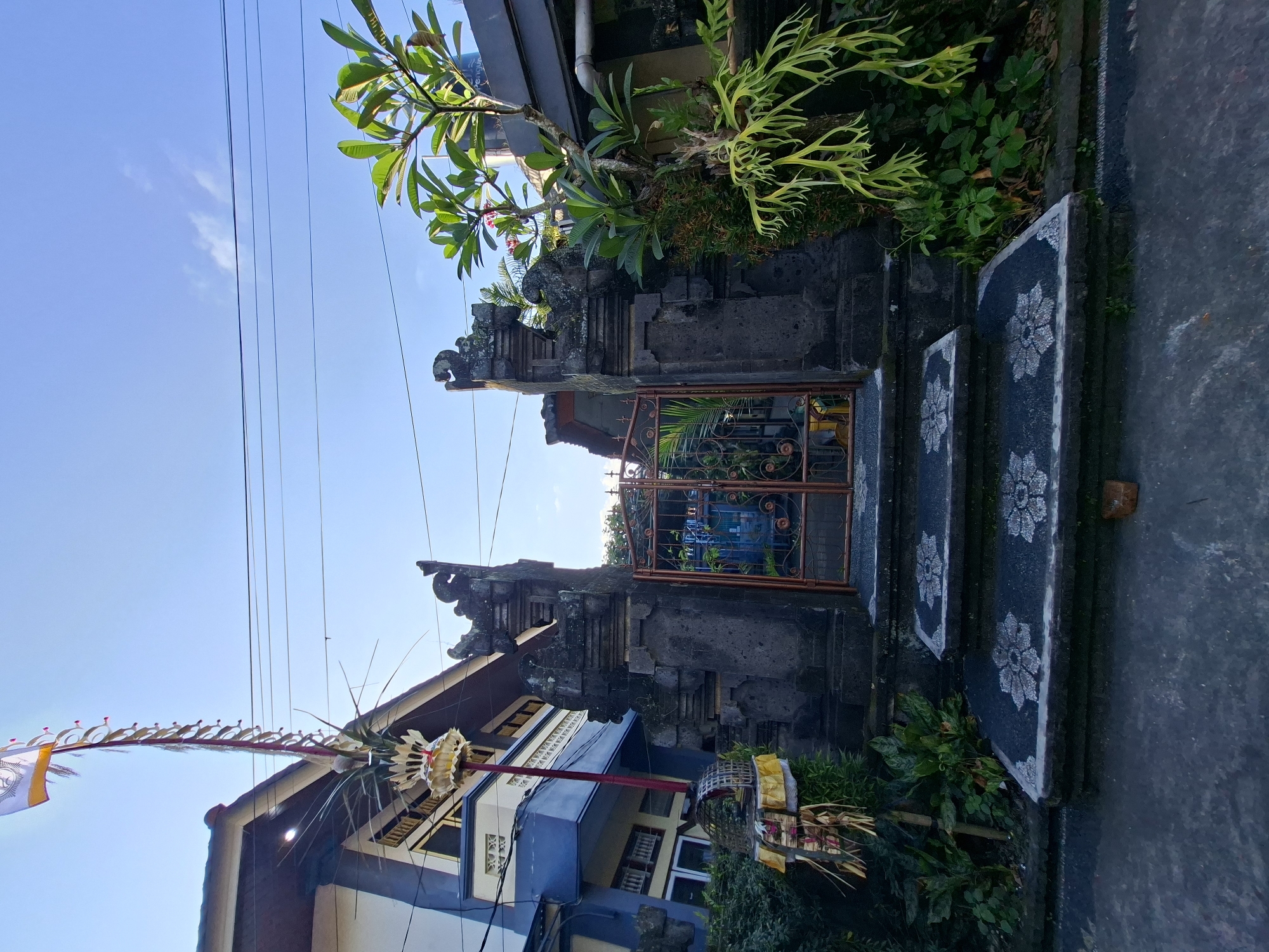 The image shows an ornate entrance gate to a building in Blahbatuh, Indonesia. The gate is made of dark stone with intricate carvings, flanked by two pillars topped with decorative figures. A set of steps with floral patterns leads up to the gate. To the left of the gate, a tall bamboo pole with a flag and offerings is erected, characteristic of Balinese ceremonies. The building behind the gate has a blue and white facade, with a prominent roofline. Lush green vegetation surrounds the entrance, and a frangipani tree with pink blossoms grows to the right of the gate. The sky is clear and blue, with power lines running across it. The scene depicts a typical residential entrance in the region, possibly decorated for a cultural event.
