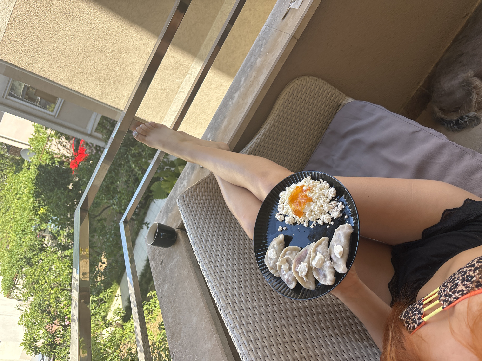 The image depicts a person relaxing on a balcony in Budva, Montenegro, enjoying a meal and a drink. The person, whose legs and torso are visible, is lying on a woven chaise lounge with a grey cushion. They are holding a black plate with a meal consisting of dumplings topped with a dollop of white cream, cottage cheese, and a spoonful of orange jam or preserve. A dark cup, possibly of coffee or tea, sits on a stone ledge to the left of the chaise lounge.
The setting appears to be outdoors on a sunny day, indicated by the strong shadows cast by the metal railing and the bright illumination of the scene. Lush green trees and foliage are visible in the background, suggesting a warm climate. The architecture in the background hints at a Mediterranean or Balkan location, consistent with the provided location context of Budva, Montenegro. The overall mood seems relaxed and leisurely, suggesting a vacation or a peaceful moment of self-care. There is no visible text in the image.