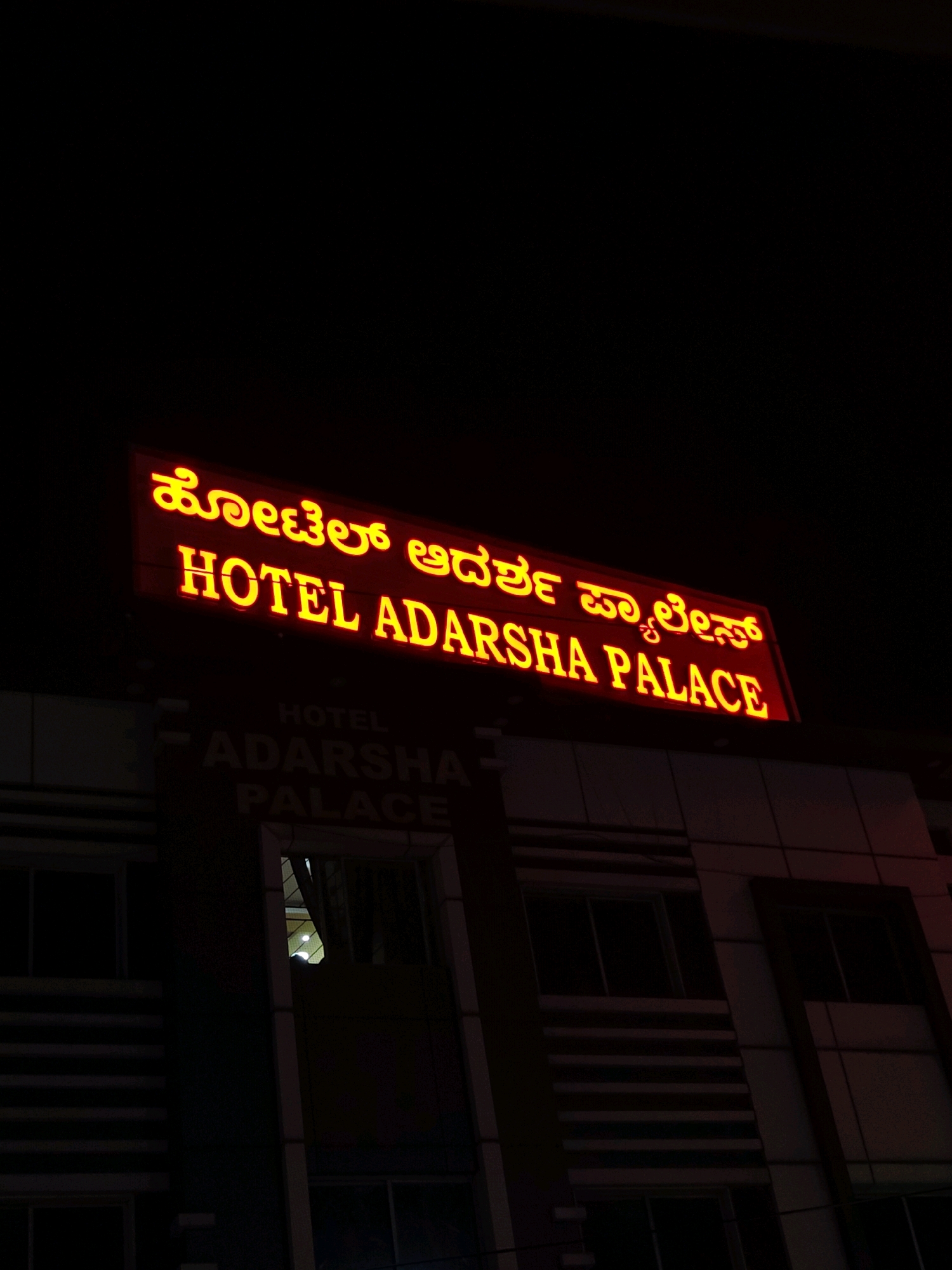 The image shows a neon sign that reads "HOTEL ADARSHA PALACE" in yellow lettering against a dark background. The sign is illuminated, and the letters are clear and distinct. Below the neon sign, there is another sign that appears to be made of brick or stone, with the words "HOTEL ADARSHA PALACE" carved into it, also illuminated. The building itself is dark, but the lit signs are prominent. The overall impression is that of a hotel at night, likely in an urban area. The building's architecture suggests it is a modern structure with multiple floors and windows. The setting appears to be a street view at night. The neon sign is positioned high on the building, indicating it is meant to be seen from a distance. The lighting creates a somewhat dramatic effect, highlighting the hotel's name and entrance. It is implied that this hotel is located in Bengaluru, India, based on the context provided.