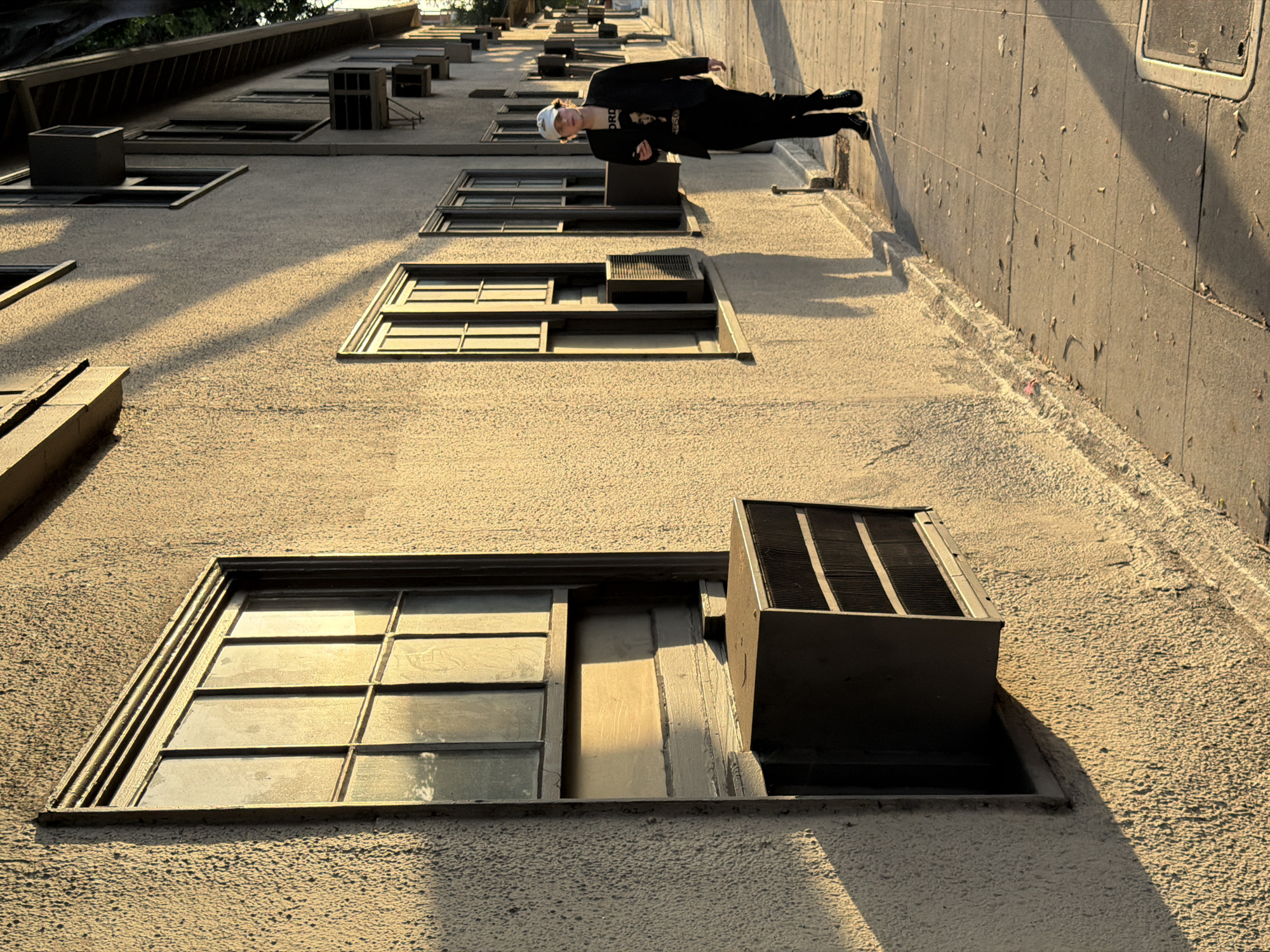 A low-angle photograph depicts a building's facade, possibly in West Hollywood, California, with a man positioned to appear as if he is leaning against the building wall. The building is a light tan color with multiple rectangular windows and air conditioning units placed in some of the window spaces. The man is wearing a black jacket and trousers with a white head covering and appears to be suspended horizontally in the frame. A shadow of the man is cast on the building. To the right, a grey concrete wall is visible, marking a potential corner of the building. The lighting suggests late afternoon, casting elongated shadows across the textured surface of the building.
