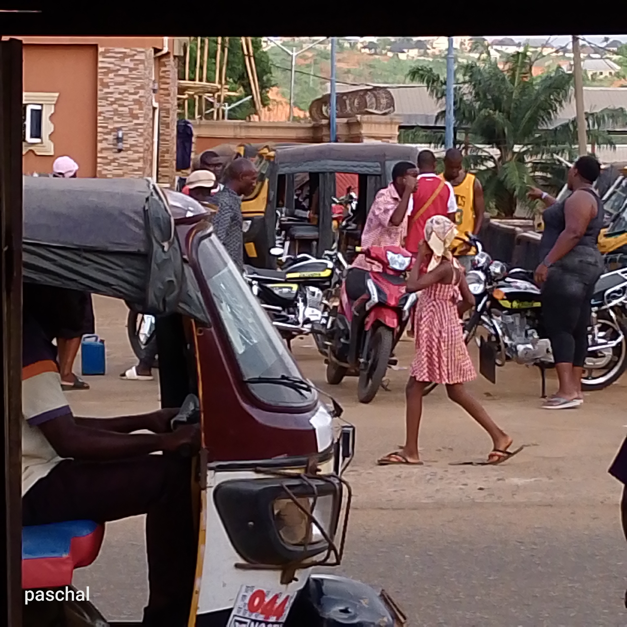 The scene is in Nsukpe, Nigeria, depicting a bustling street with various individuals and vehicles. A tricycle (auto-rickshaw), with the driver visible, occupies the foreground. In the background, a group of people stand near motorcycles, one of whom is gesturing. A young girl wearing a dress and headscarf is walking. There are buildings and trees visible in the background.