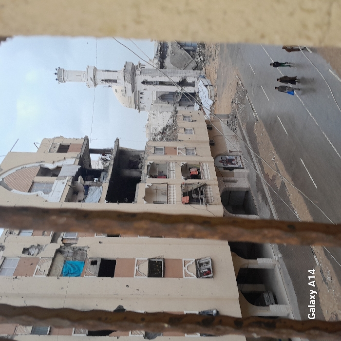 The image displays a war-damaged urban street scene in Khan Yunis, Palestinian Territory, viewed through vertical brown bars or beams. In the mid-ground, a multi-story building with a beige facade exhibits extensive structural damage, including collapsed sections on upper floors, visible rebar, numerous impact holes, and shattered windows. A blue cloth hangs from one window, and laundry is visible on some balconies. The ground floor features arched openings, with one revealing a parked light-colored vehicle.
To the right of this building, a mosque with a white dome and a tall, slender minaret topped with a crenellated structure is visible, also showing significant damage to its facade and surrounding structures, which are reduced to rubble.
In the foreground, two horizontal wires or cables stretch across the scene. Below them, a wet, dark asphalt road with white lane markings shows reflections of the overcast sky. Four individuals are walking across the road from left to right; one carries a blue bag. The sky above is uniformly grey and overcast. A "Galaxy A14" watermark is present in the bottom-left corner.