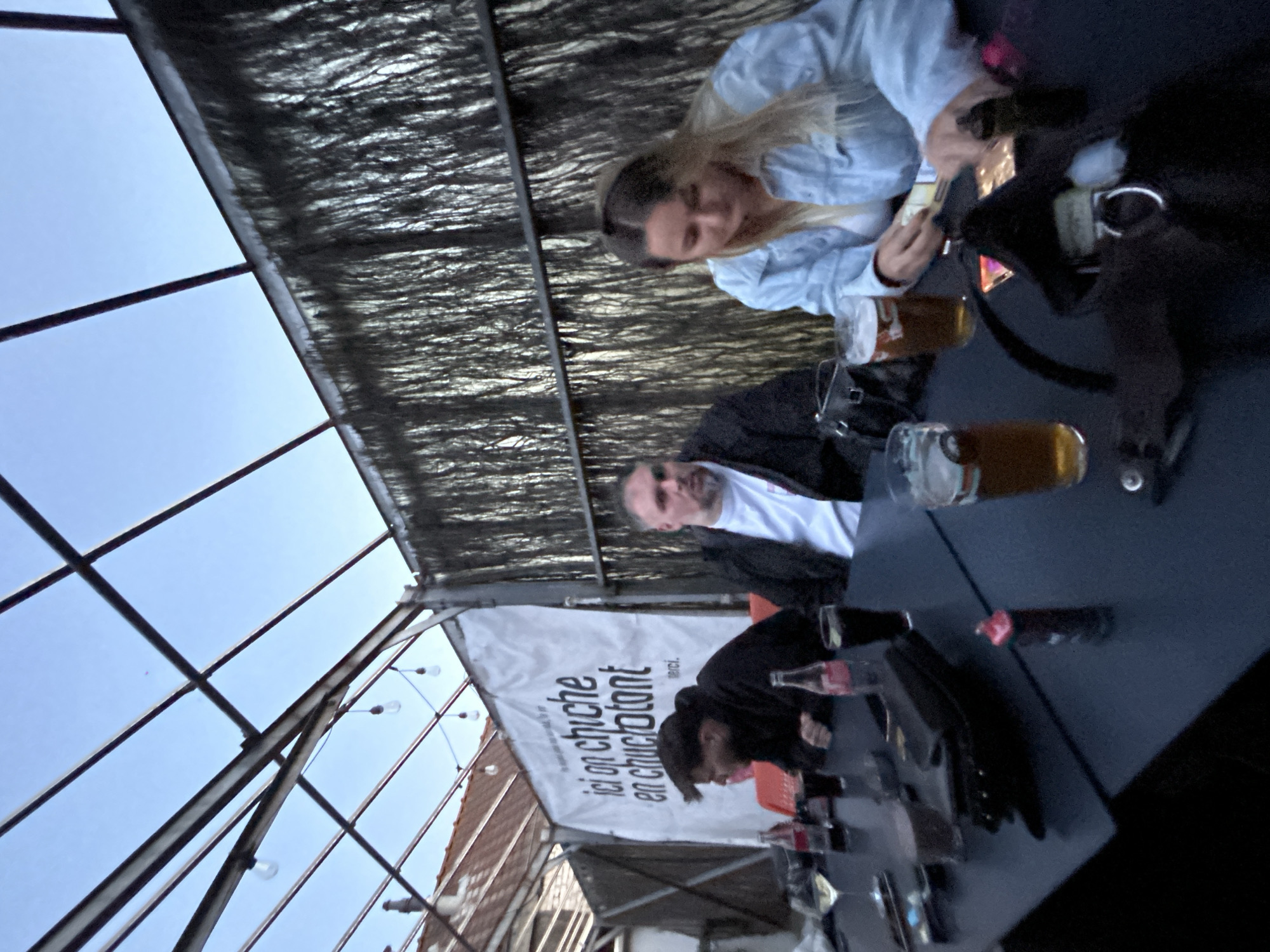 The image captures a casual gathering at an outdoor or semi-outdoor seating area, likely a covered terrace, on a clear day. A bright blue sky is visible through a dark metal framework and above a textured, dark canopy that provides shade. Bare lightbulbs are strung along the framework, suggesting decorative lighting or preparations for evening use.
Three people are seated around a dark table. To the right, a woman with blonde hair, wearing a light blue jacket, is engrossed in an activity at the table or within her bag. Two glasses of an amber beverage, likely beer, are positioned near her. Directly opposite, a man with short dark hair and a beard, dressed in a black jacket over a white t-shirt, looks towards the camera. He also has a glass of the amber drink in front of him. Further to the left, partially obscured, another person in a black hoodie is leaning forward, with two Coca-Cola bottles visible nearby.
A white banner with black text is prominently displayed in the background. The text reads: "ici on chiche en chuculatant incl. merci." This phrase, possibly a playful local slogan or idiom related to a challenge or chocolate, points to a French-speaking environment, consistent with the location context of Hem, France. The scene depicts a relaxed social interaction over drinks.