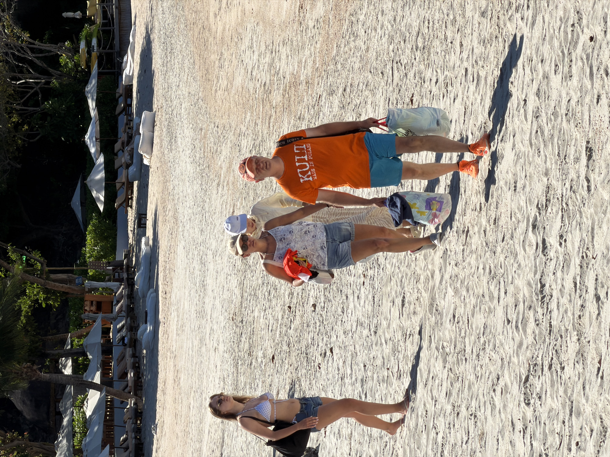 Four people are on a sandy beach near a row of lounge chairs and umbrellas. The individuals appear to be a mix of adults, with two men and two women.
The first man from the top is wearing an orange t-shirt with the text "KULT MADE IN POLAND" and blue shorts. He is holding a white bag and wearing orange sneakers. Next to him is a woman in a white top with a patterned design and denim shorts, also wearing a blue cap. She is holding a red towel and a small white bag.
Below them, a woman is lying on the sand wearing a white polka dot bikini and denim shorts. She is wearing sunglasses and a black bag is next to her. The fourth person's lower body is visible in the upper left quadrant of the image, wearing a light-colored garment and resting on the sand.
The scene is set on a beach with sand, vegetation, and lounge chairs. The lighting suggests it is daytime, likely late afternoon due to the long shadows cast on the sand. The environment appears to be tropical or subtropical, possibly in Baan Plai Laem, Thailand, as indicated by the palm trees and lush greenery in the background. The people seem relaxed and are enjoying their time at the beach.