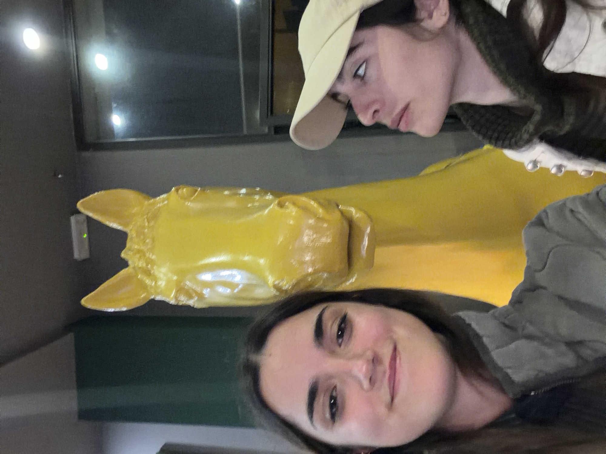 Two women are posing with a large yellow horse sculpture. One woman is at the bottom of the frame, smiling at the camera. She has dark hair and is wearing a grey jacket. The other woman is at the top of the frame, wearing a beige cap and looking to the side. Behind the sculpture, there is a dark wall with a window reflecting light. This photo could have been taken near the Leonardo da Vinci Horse Sculpture at the Ippodromo SNAI San Siro racecourse in Milan, Italy.